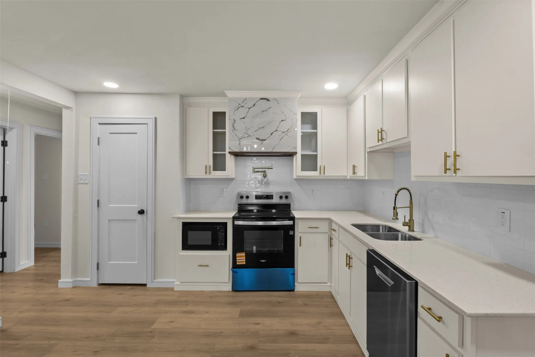 Kitchen featuring white cabinetry, light-toned countertops, a stainless steel double basin sink with a gold-finish faucet, and wood-finish flooring