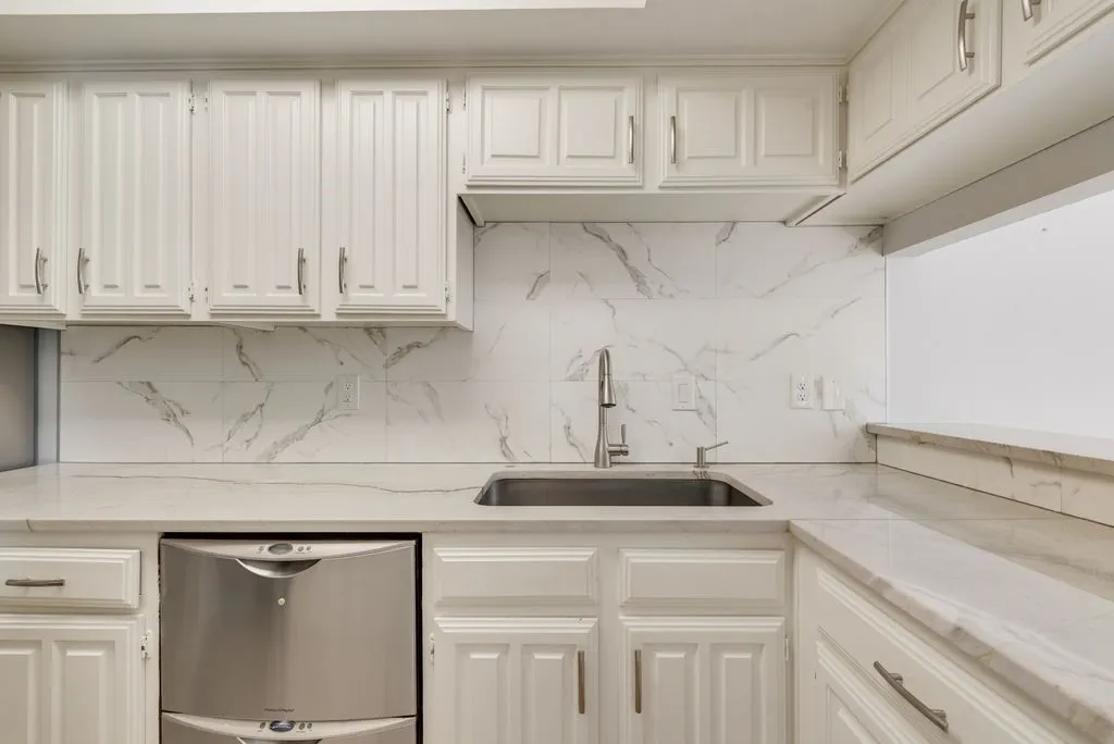 White cabinetry with brushed nickel hardware, stainless steel sink, pull-down faucet, white stone countertops, and a white tile backsplash
