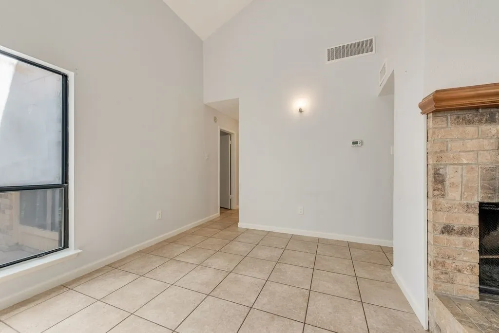 Vaulted ceiling, large window, tile flooring, brick fireplace with wood mantel, wall-mounted light fixture