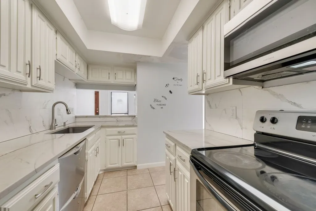 Galley kitchen featuring white cabinetry with brushed nickel hardware, tile countertops, stainless steel appliances including double wall ovens, and a window overlooking the front entrance