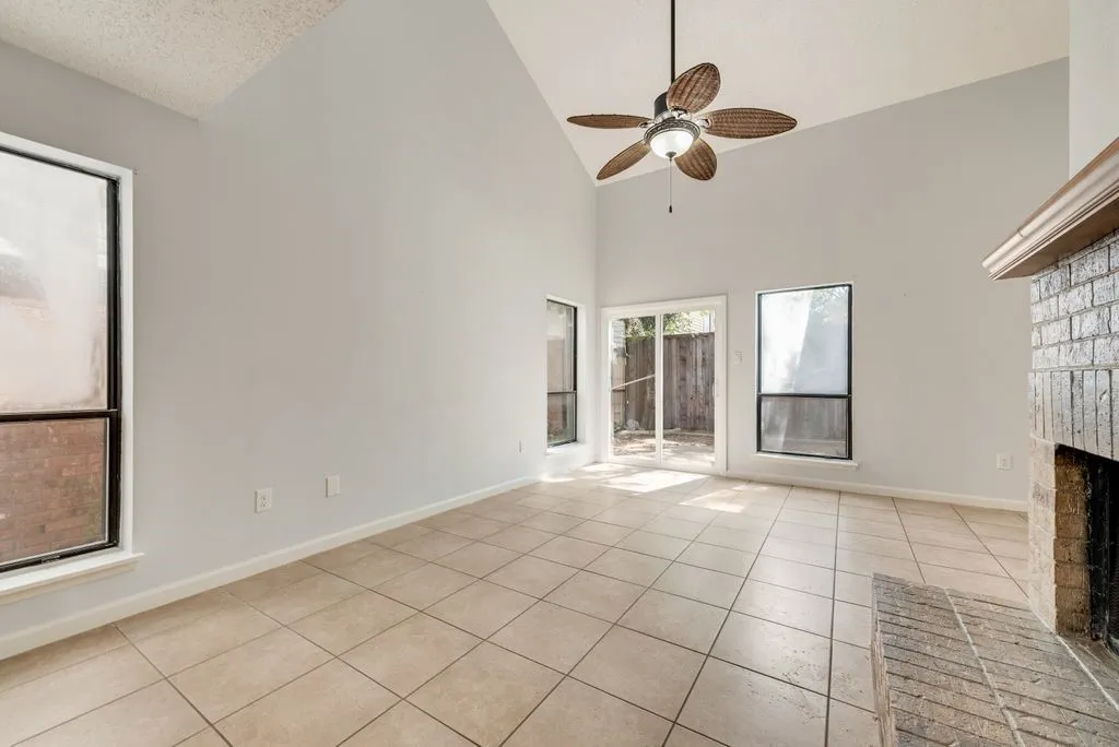 Vaulted ceiling with ceiling fan, tile flooring, brick fireplace with wood mantel, multiple windows, and a sliding glass door