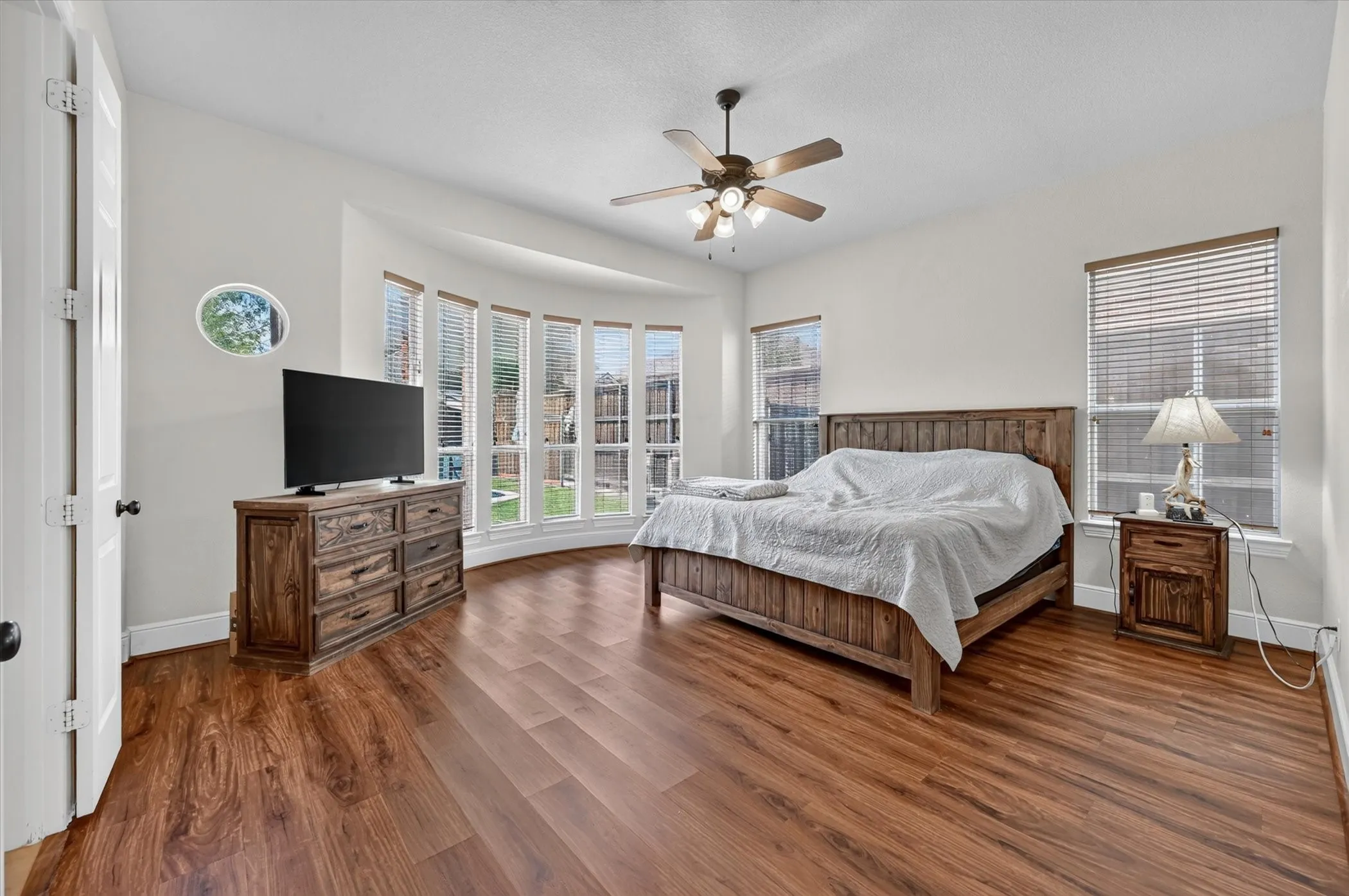 Wood-finish flooring, ceiling fan with integrated lighting, bay window feature, multiple windows with blinds, and a circular accent window