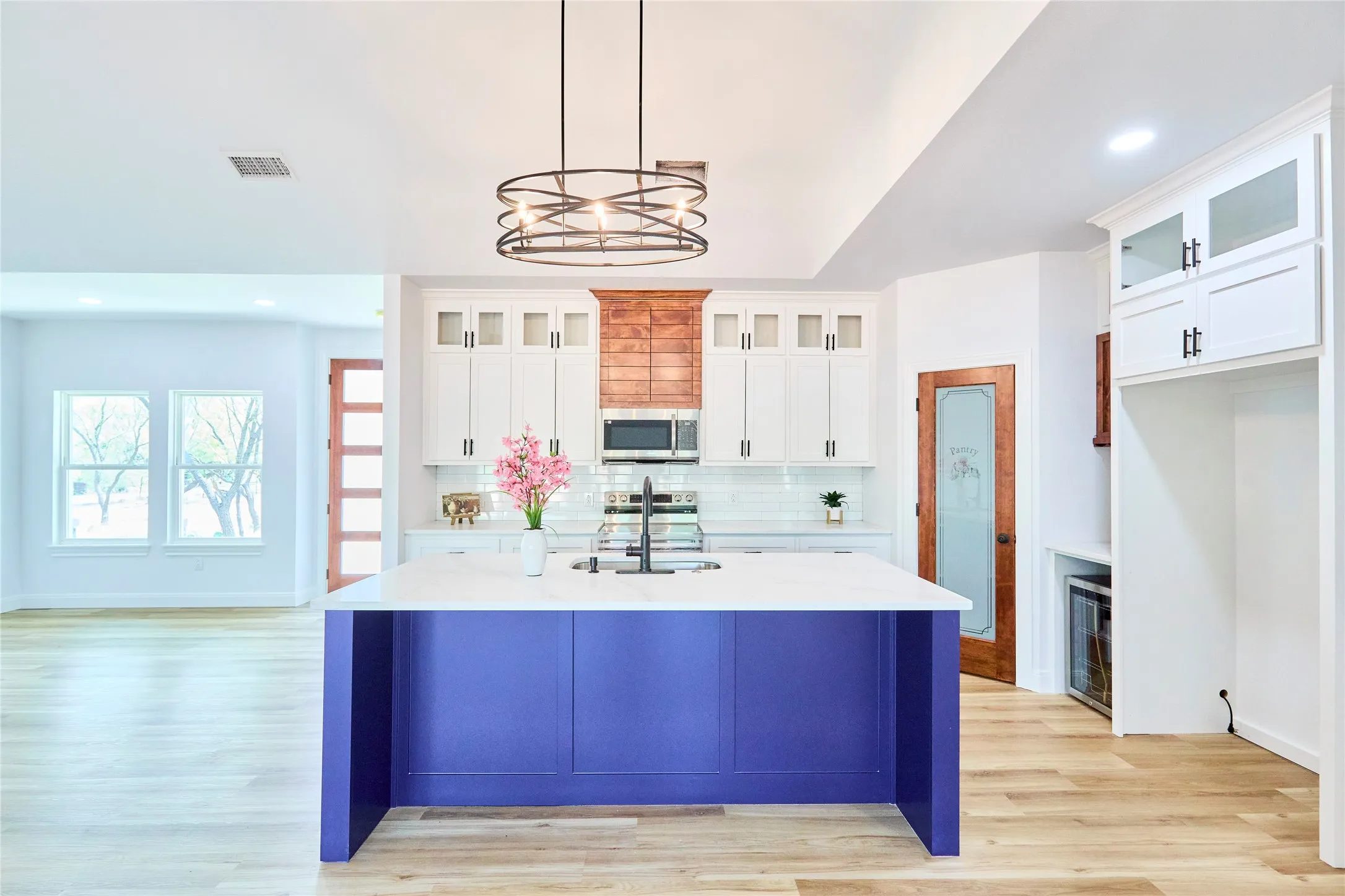 Modern kitchen featuring a central island with a built-in sink, white cabinetry with black hardware, wood-finish flooring, and a contemporary light fixture