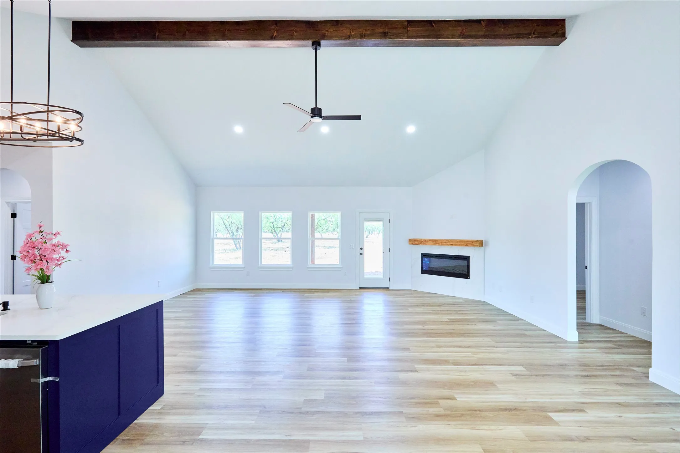 Vaulted ceiling with exposed wood beam, wood-finish flooring, electric fireplace with wood mantel, multiple windows, and arched doorway