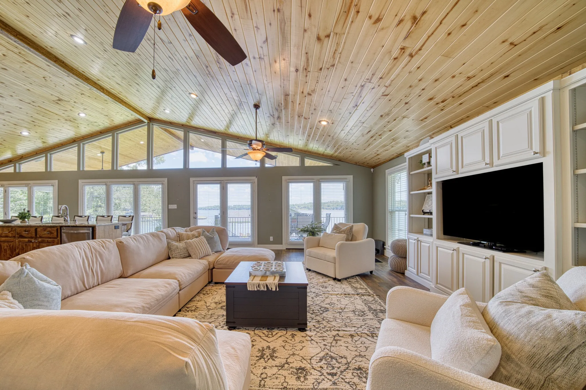 Vaulted wood-paneled ceiling with exposed beams, built-in entertainment center, multiple ceiling fans, and a wall of windows and glass panel doors