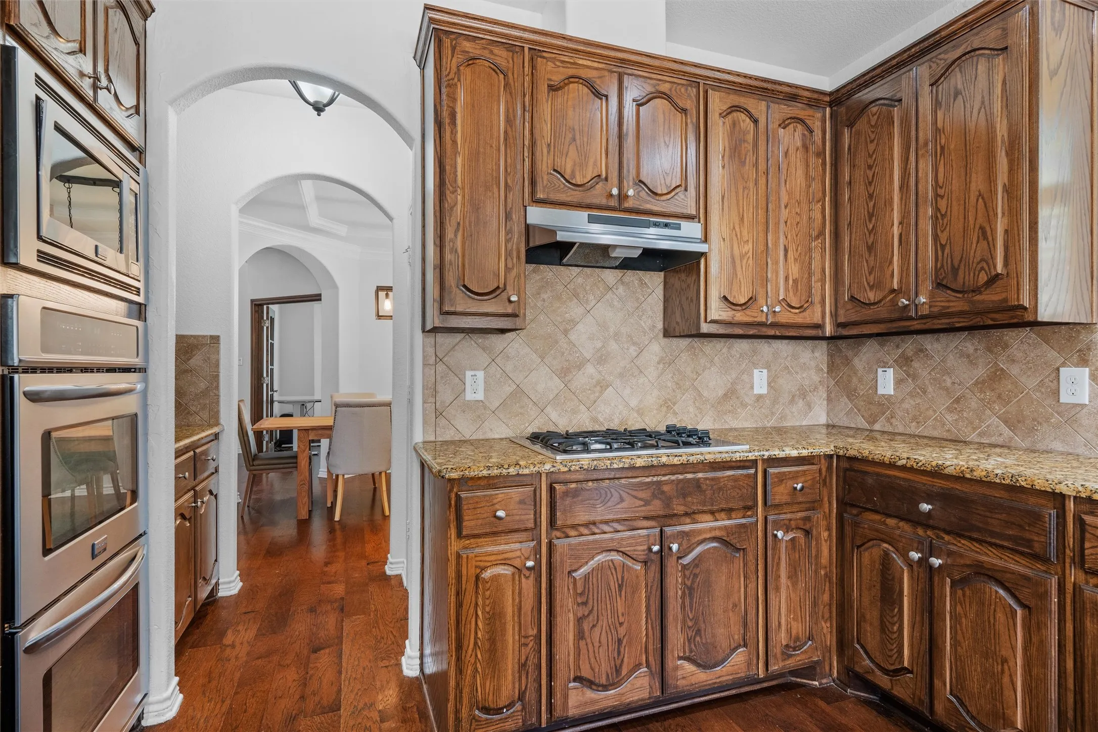 Kitchen featuring rich wood cabinetry, stainless steel double ovens, a gas cooktop, natural stone countertops, and a tiled backsplash