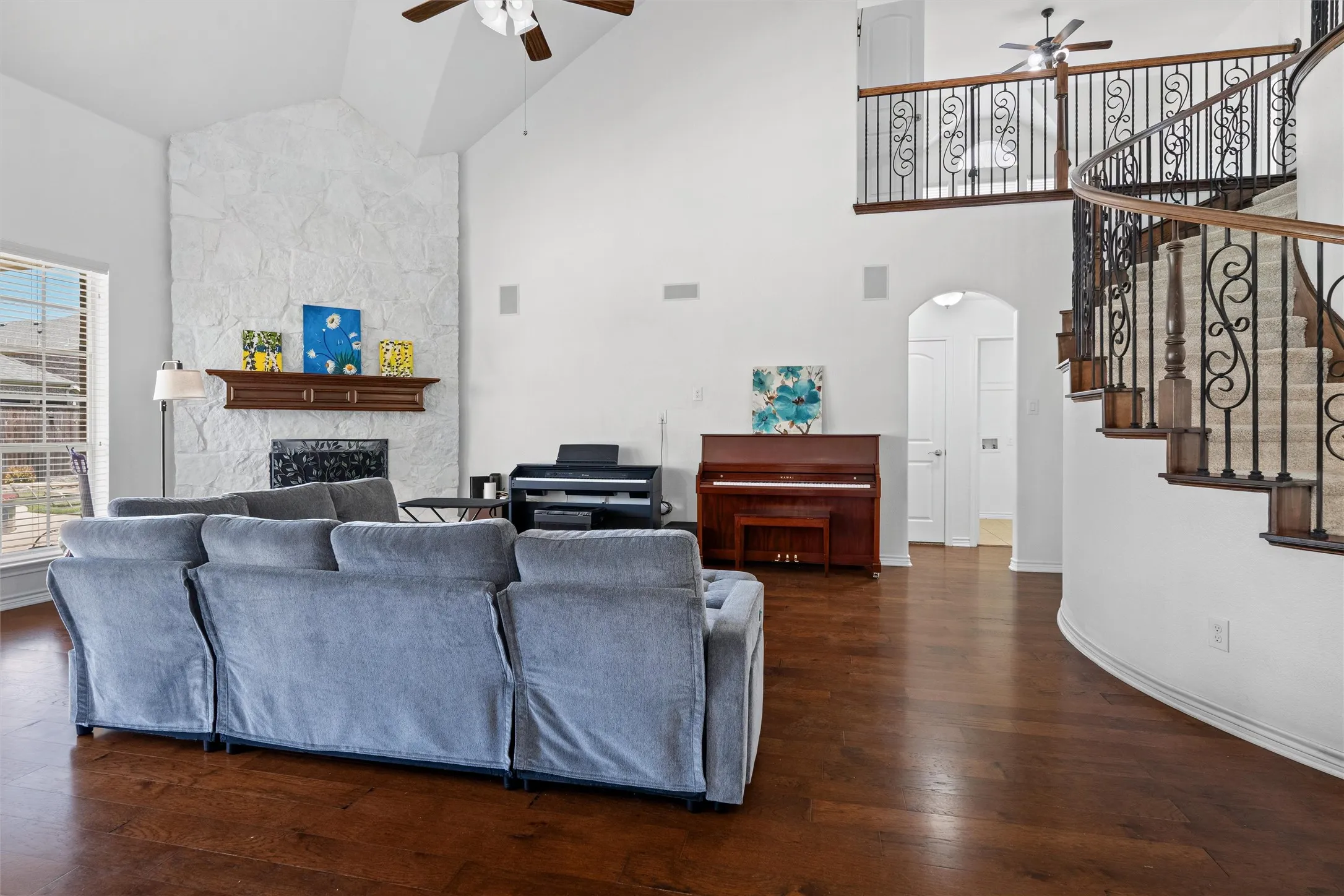 Soaring vaulted ceiling, wood-finish flooring, stone-clad fireplace with mantel, and a curved staircase with wrought iron balusters