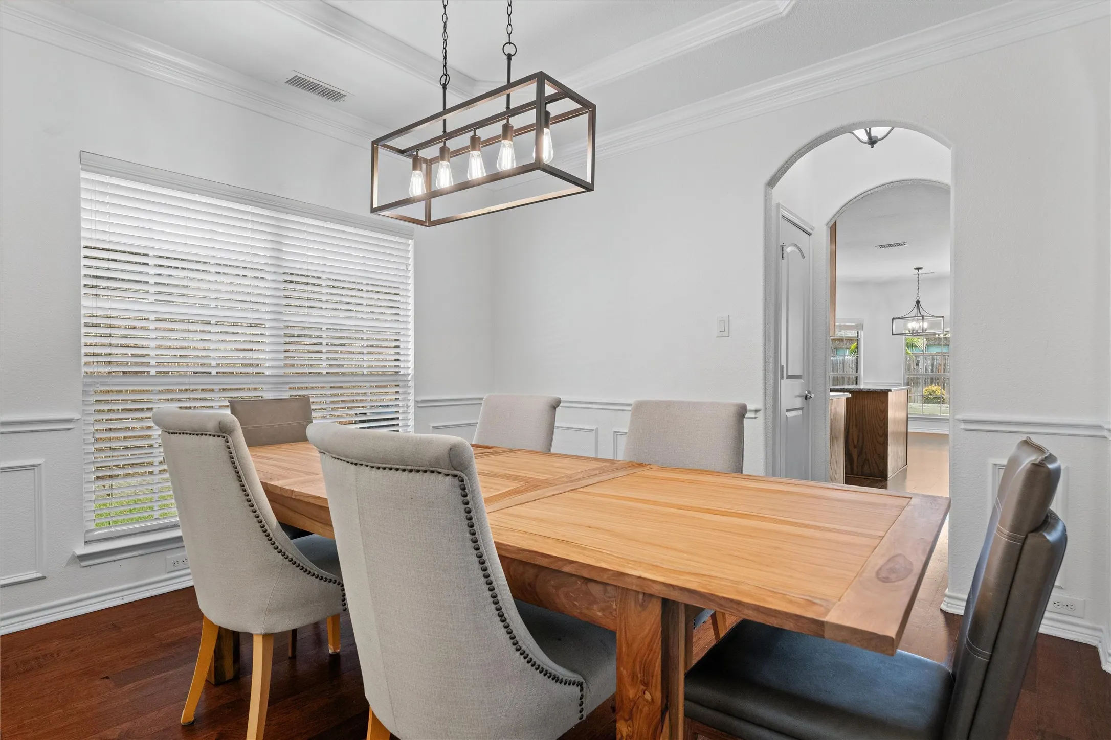Wood-finish flooring, crown molding, wainscoting, large window with blinds, and an arched doorway