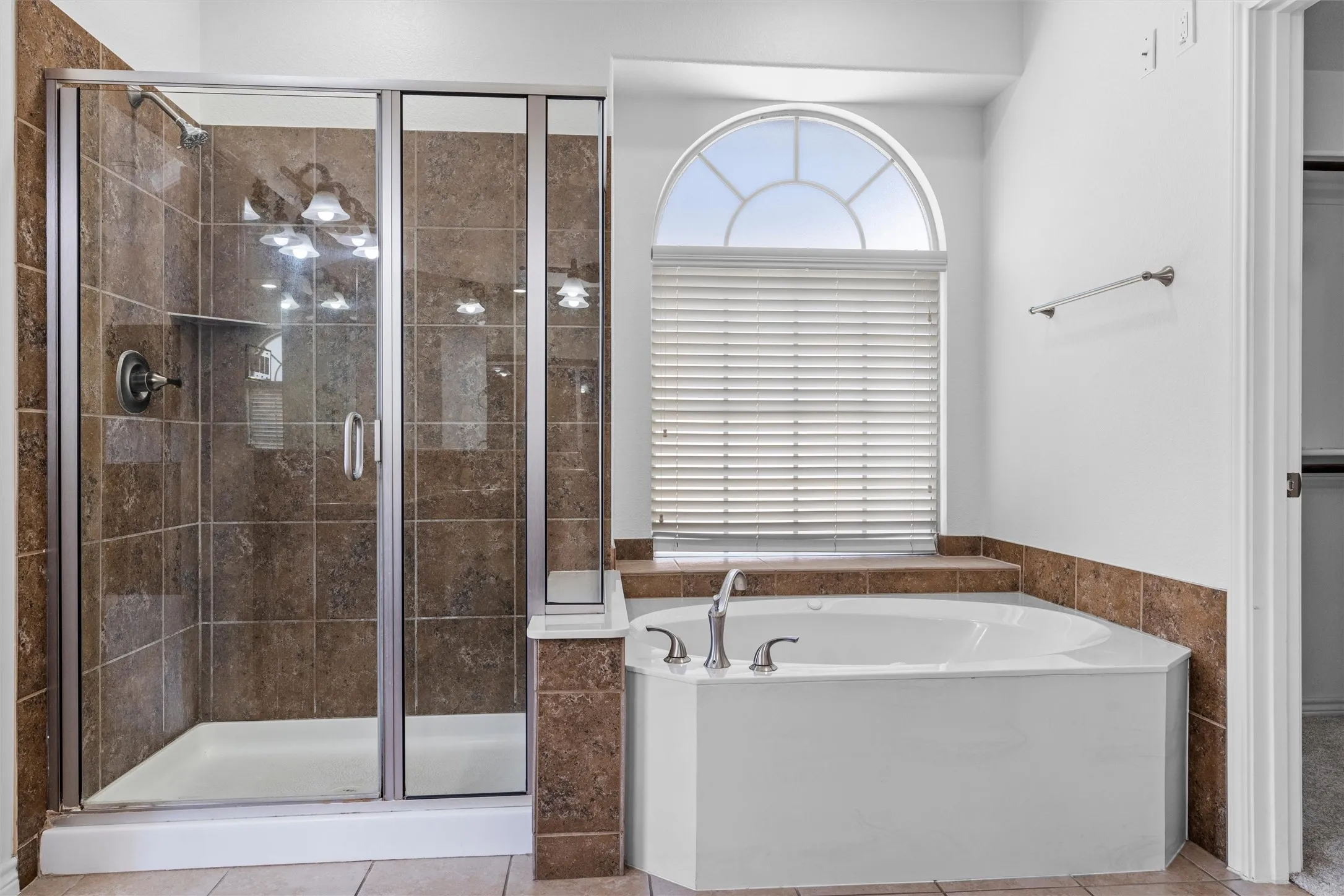 Tiled walk-in shower with a glass enclosure, corner soaking tub with deck-mounted fixtures, and an arched window with blinds