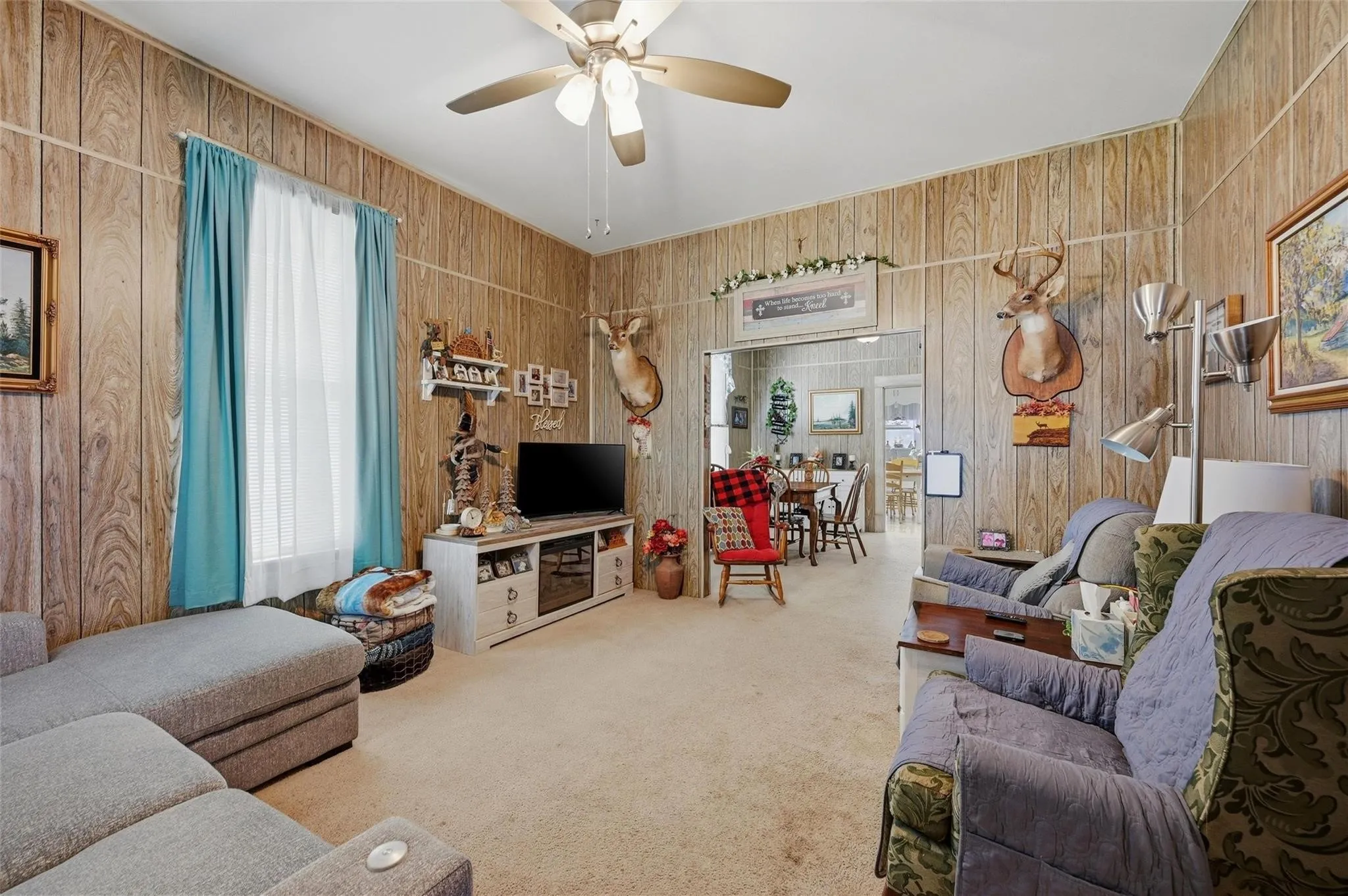 Carpeted living area with wood paneling, ceiling fan, and a large window