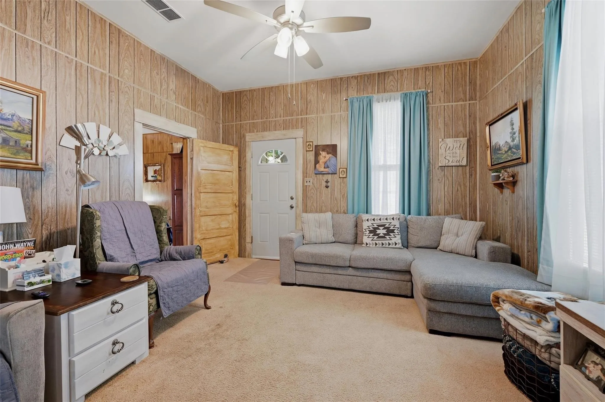 Wood-finish vertical wall paneling, light-toned carpeting, white ceiling fan with integrated lighting, white paneled entry door with decorative glass, and a large window with light blue drapes