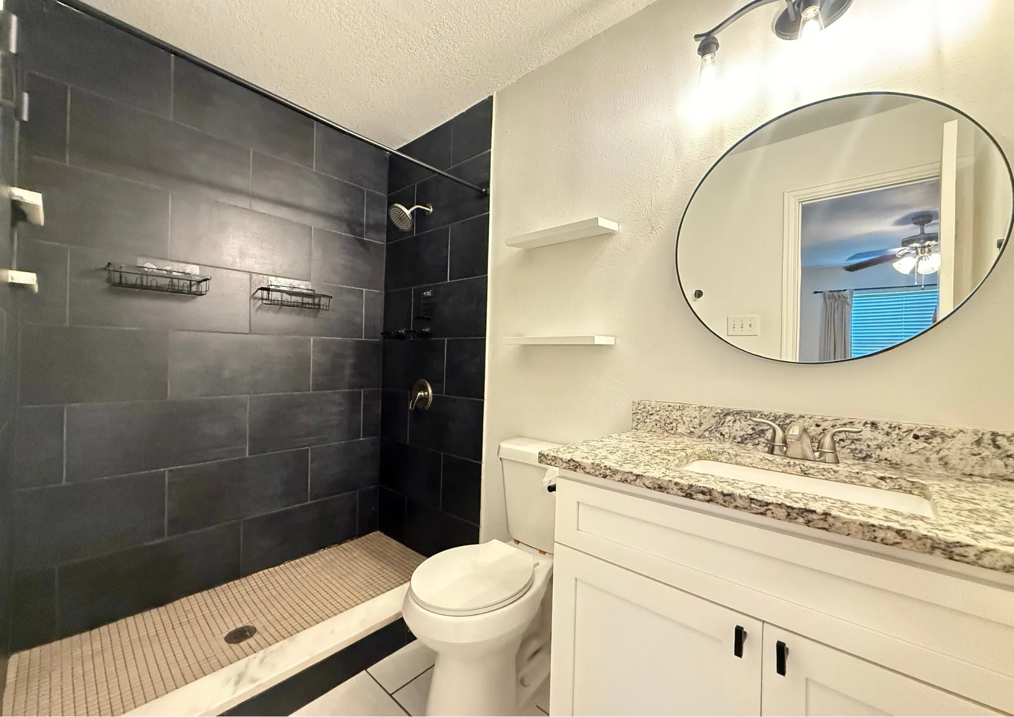 Walk-in shower with dark tile surround, white vanity with stone-look countertop, integrated sink, and matte black hardware