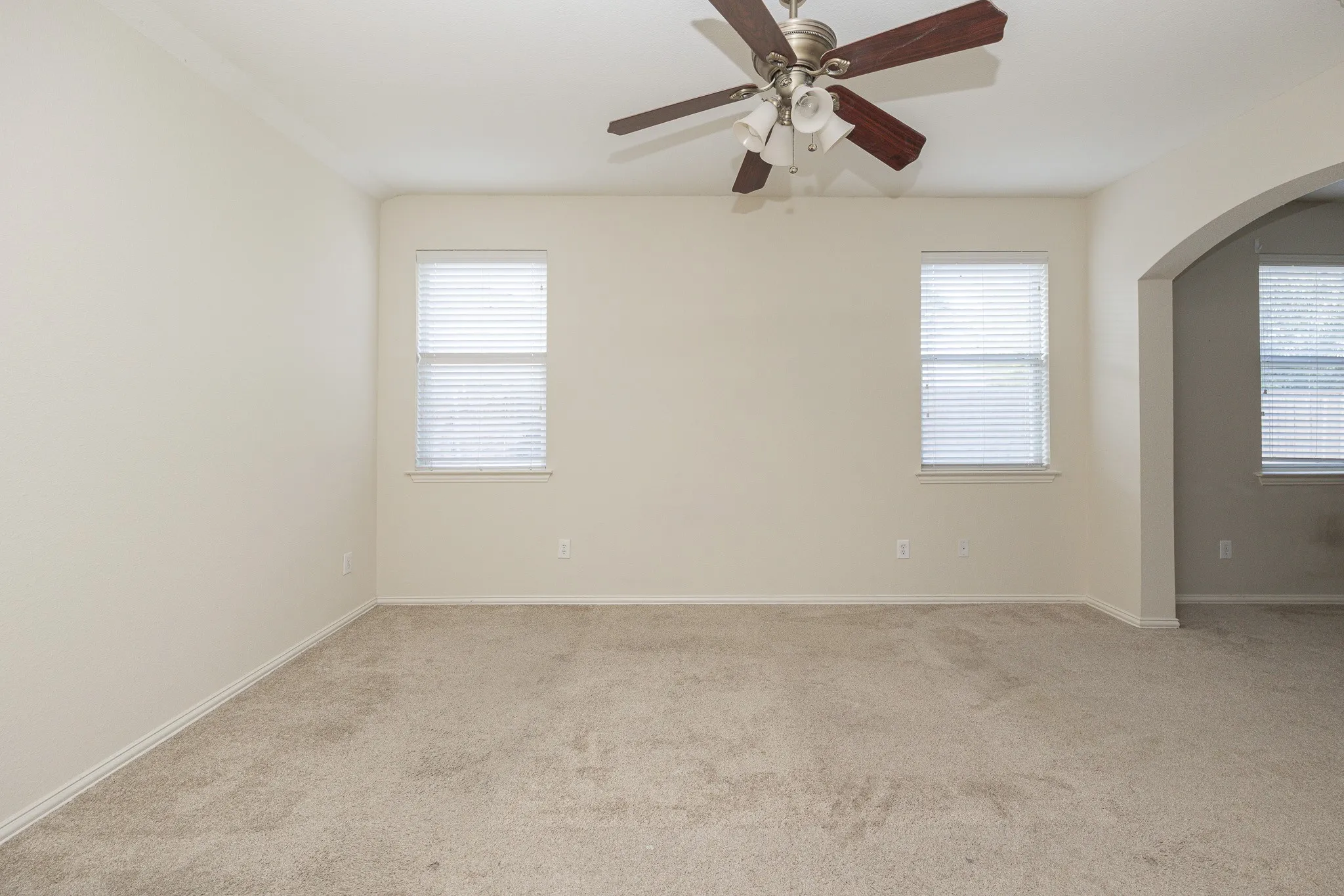 Neutral toned walls, light carpeting, two windows with blinds, and a ceiling fan with light fixture