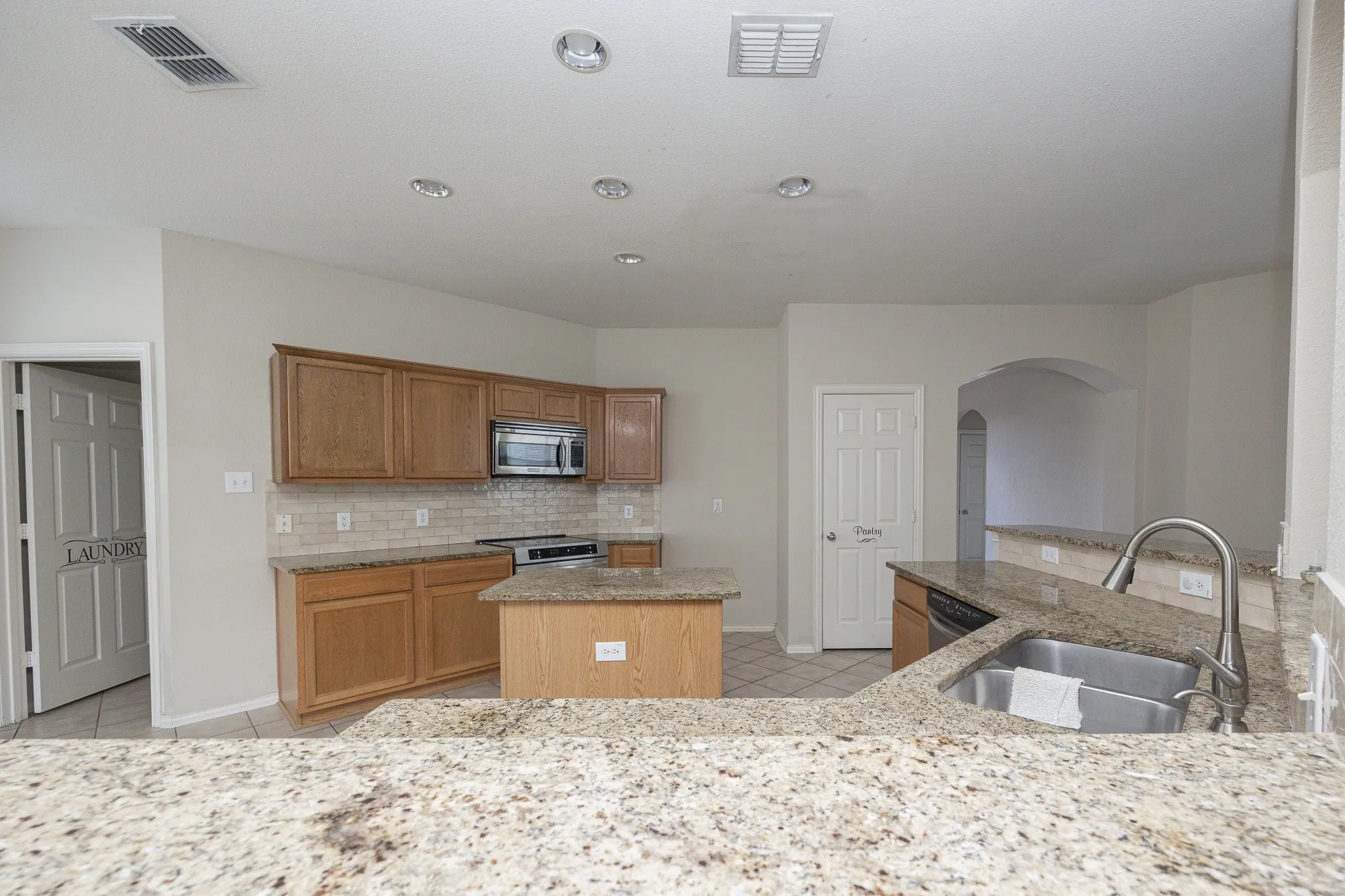Spacious kitchen featuring warm wood cabinetry, stainless steel appliances including a built-in microwave, a large center island with seating, speckled stone countertops, and a tile backsplash