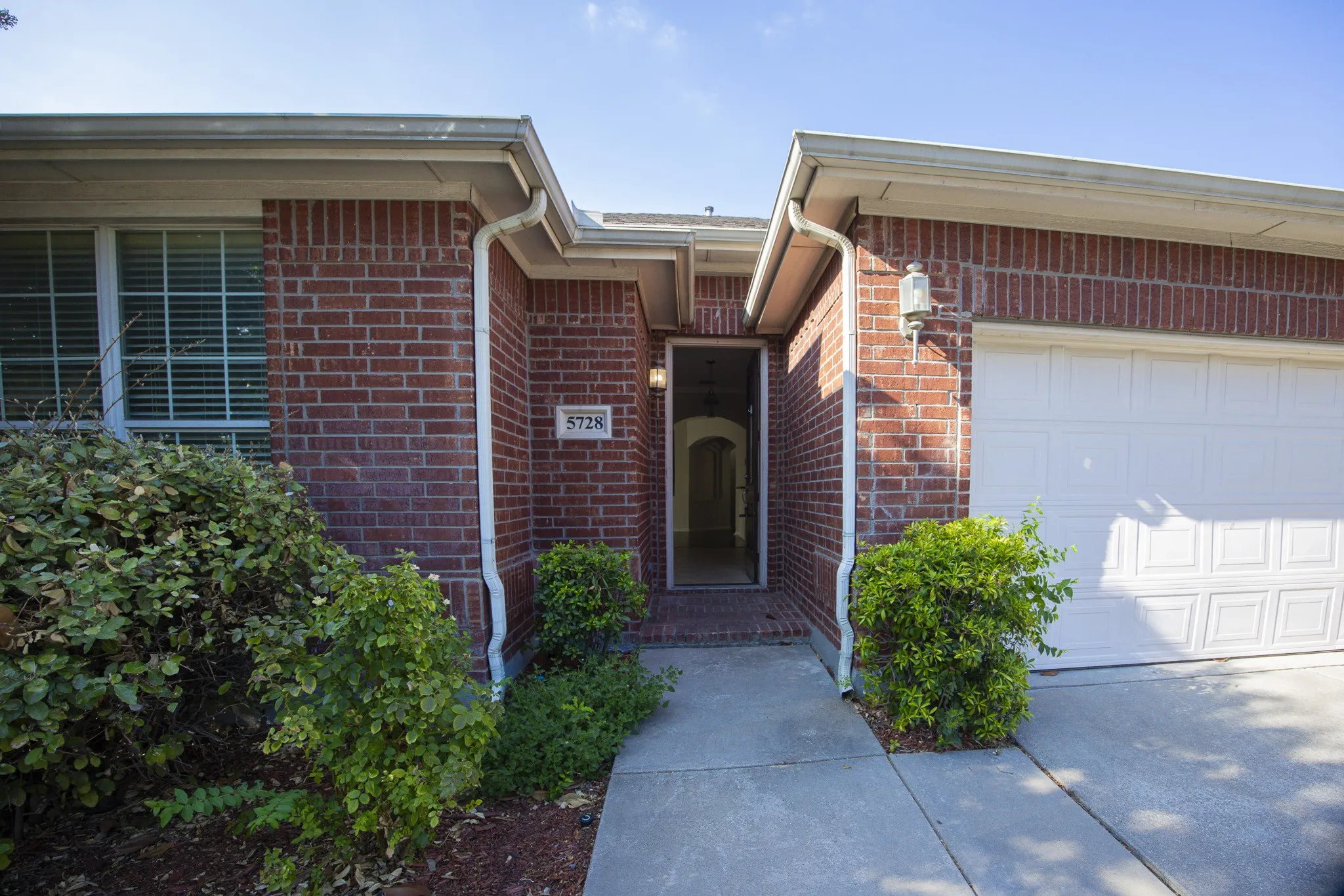 Brick exterior with an attached garage, featuring a concrete walkway, mature landscaping, and an exterior light fixture