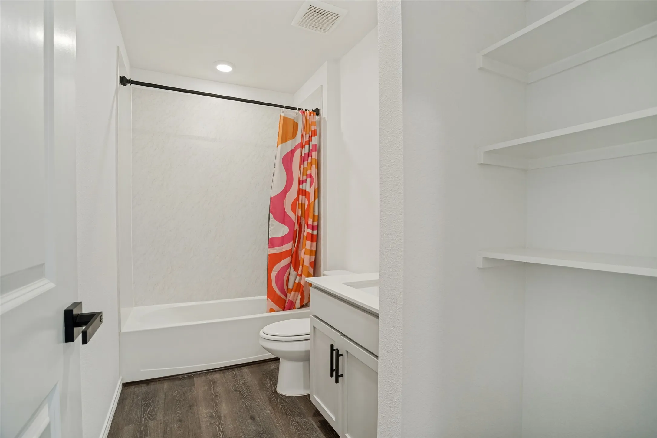 Shower-tub combination with a white textured surround, vanity with a white countertop and dark hardware, wood-finish flooring, and built-in shelving