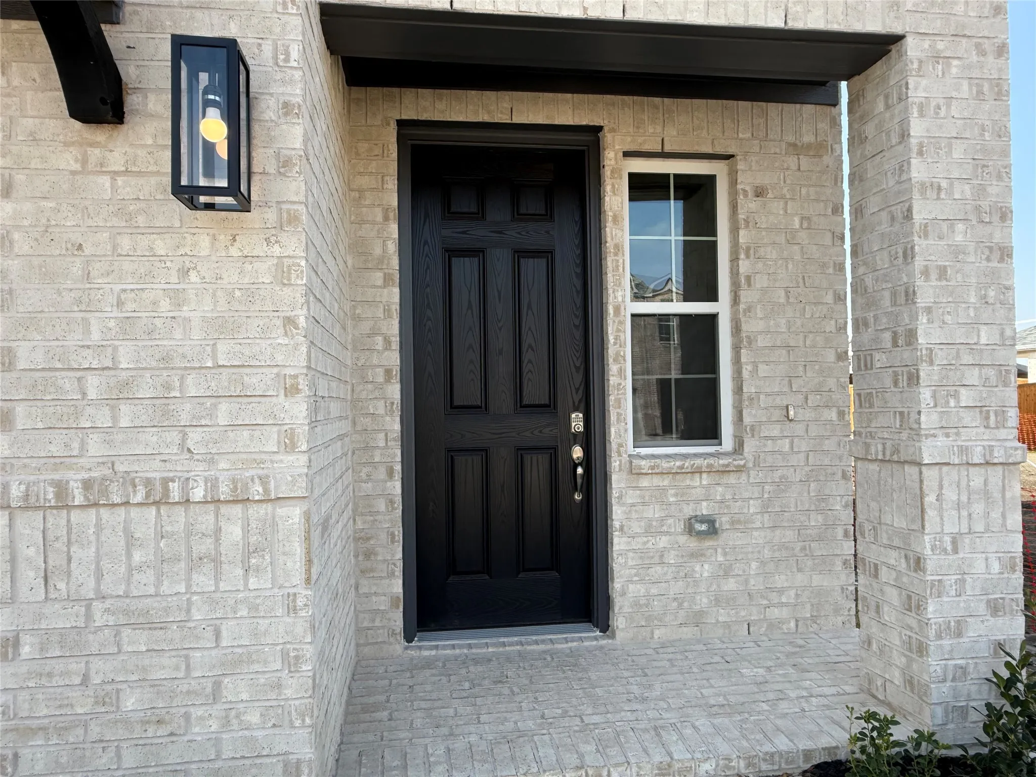 Light-toned brick exterior with a dark-paneled entry door, a single-pane window with grid inserts, an exterior wall lantern, and a covered porch