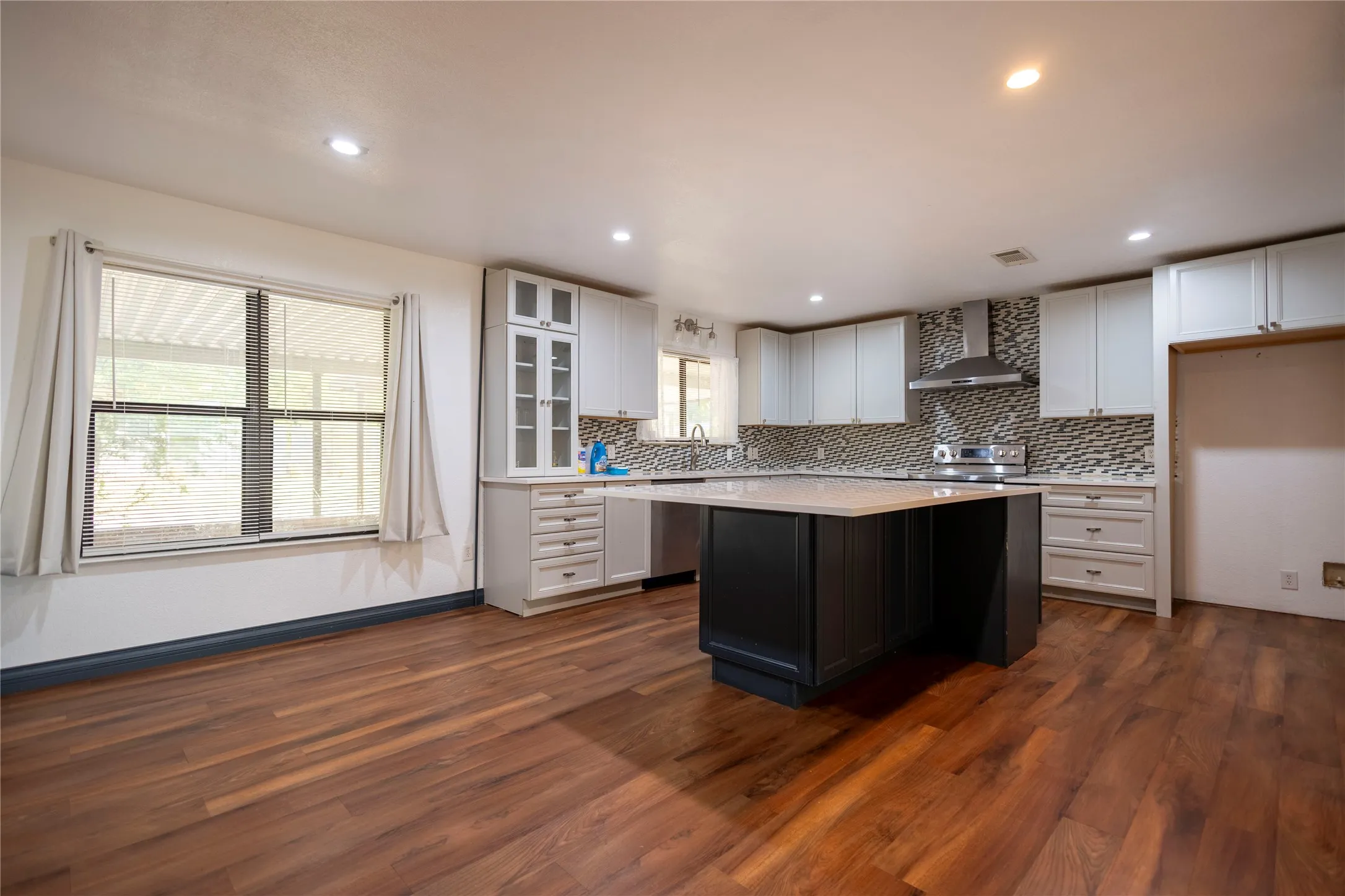 Wood-finish flooring, recessed lighting, kitchen island with solid surface countertop, stainless steel appliances, and a mosaic tile backsplash