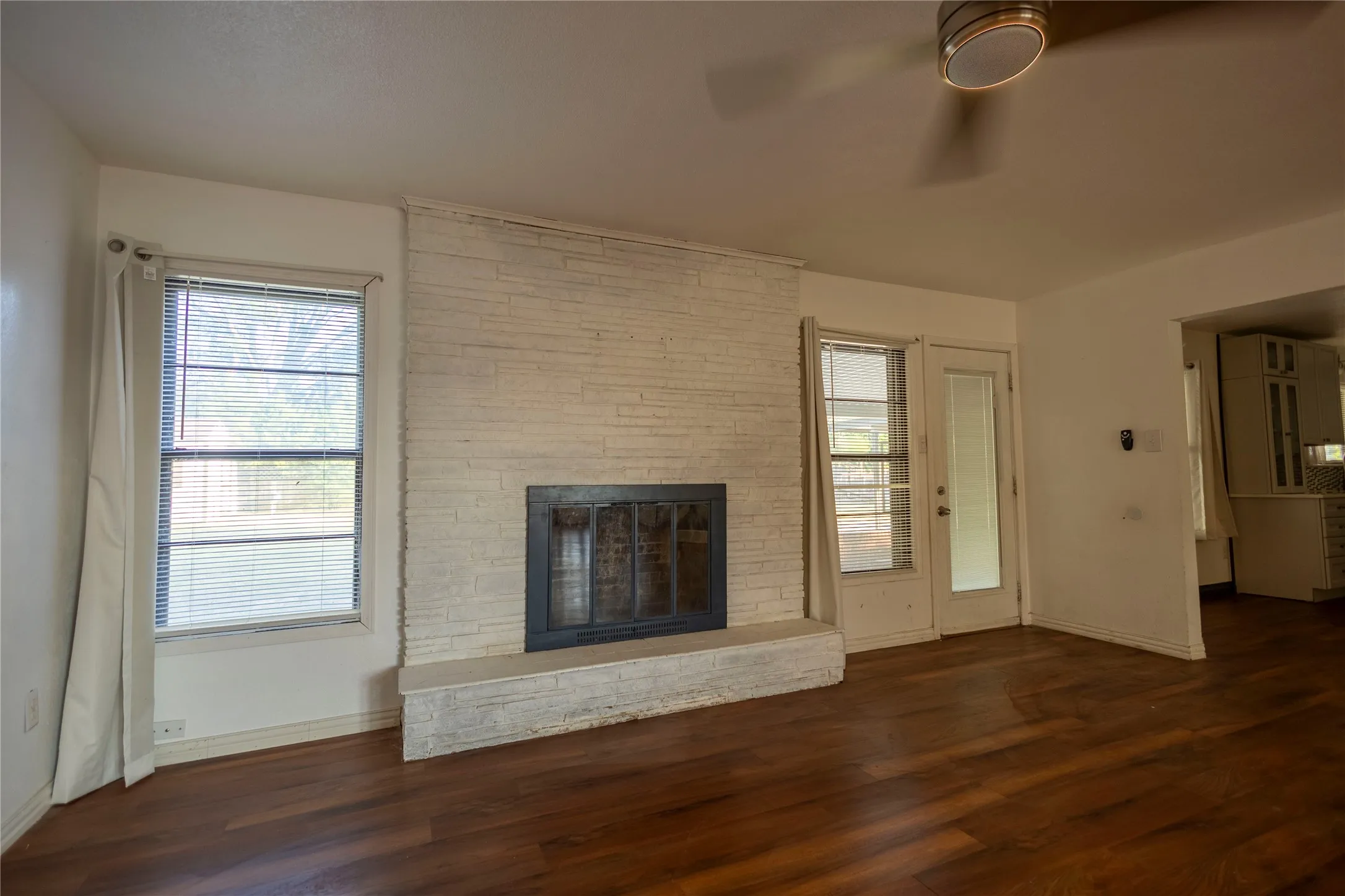 Wood-finish flooring, white brick fireplace, multiple windows, and a ceiling fan
