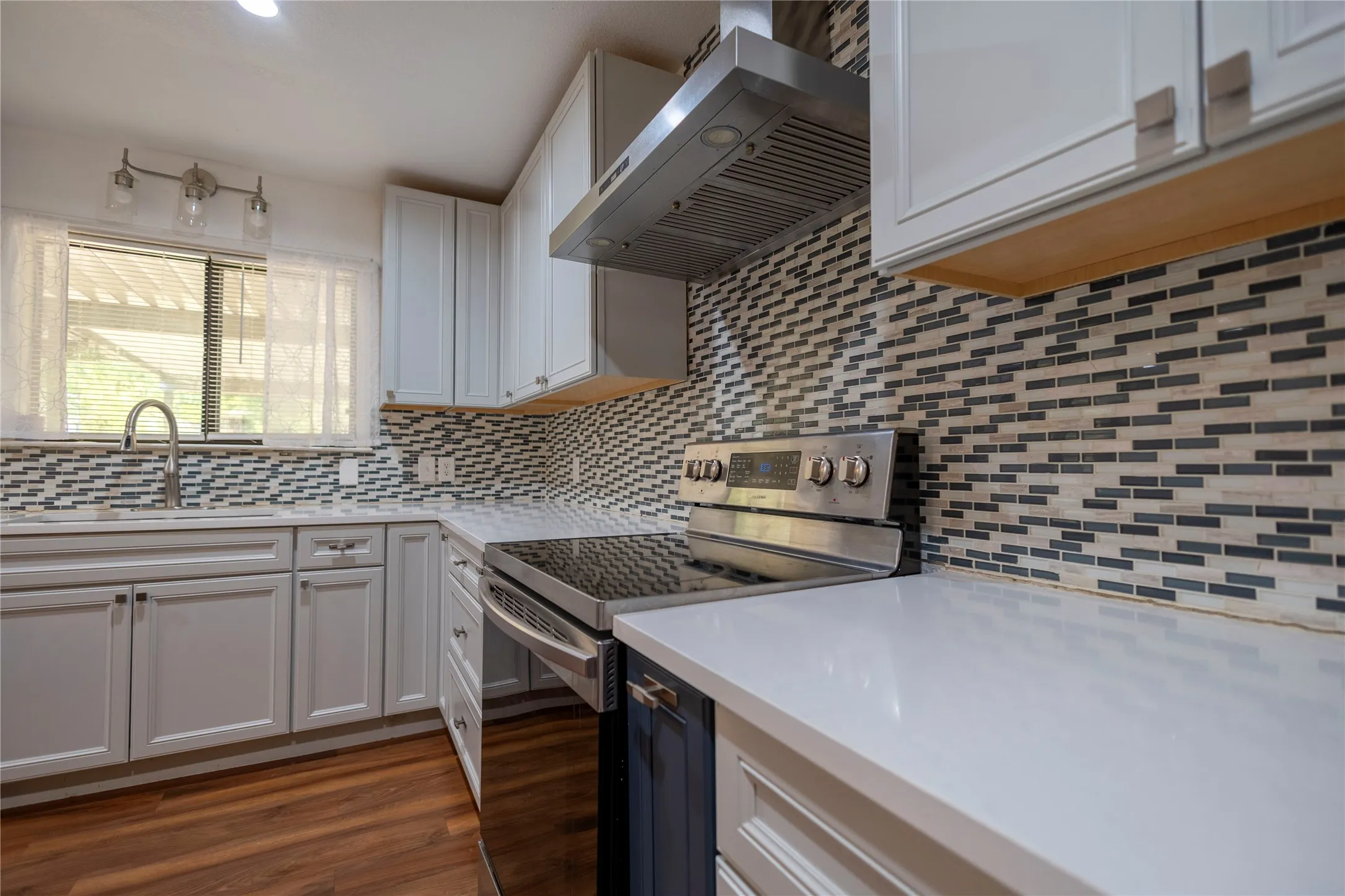 White cabinetry with modern hardware, stainless steel appliances, solid surface countertops, mosaic tile backsplash, and wood-finish flooring