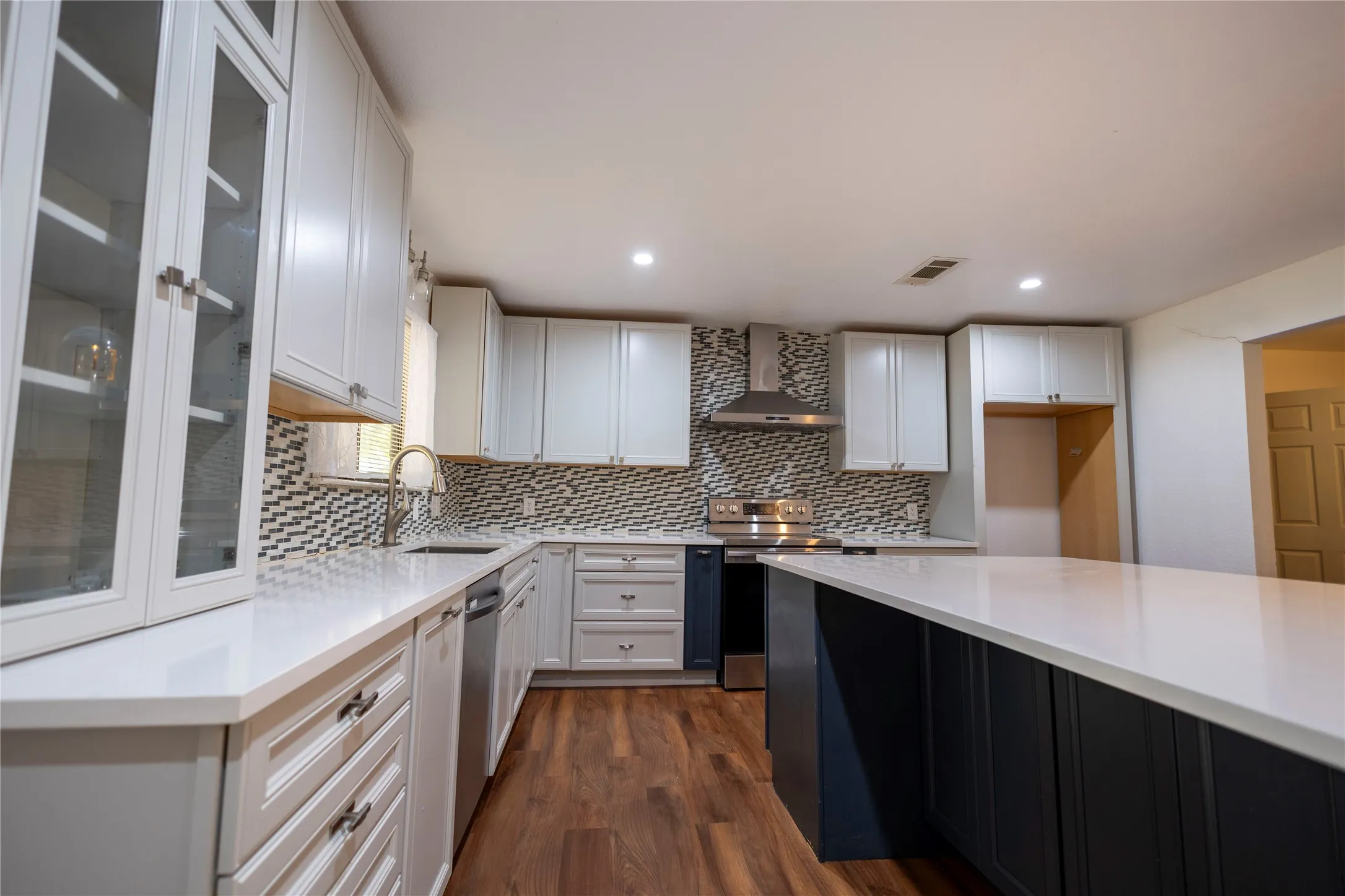 Kitchen featuring white shaker cabinetry, an expansive island with white countertops, wood-finish flooring, a stainless steel range with hood, and a decorative tile backsplash