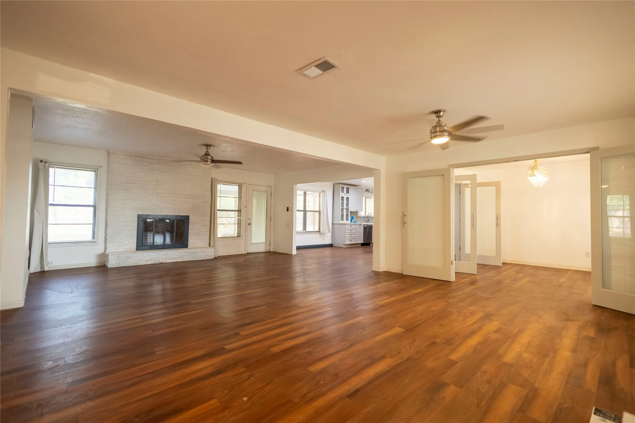 Expansive living area with wood-finish flooring, a white brick fireplace, multiple windows, and dual ceiling fans