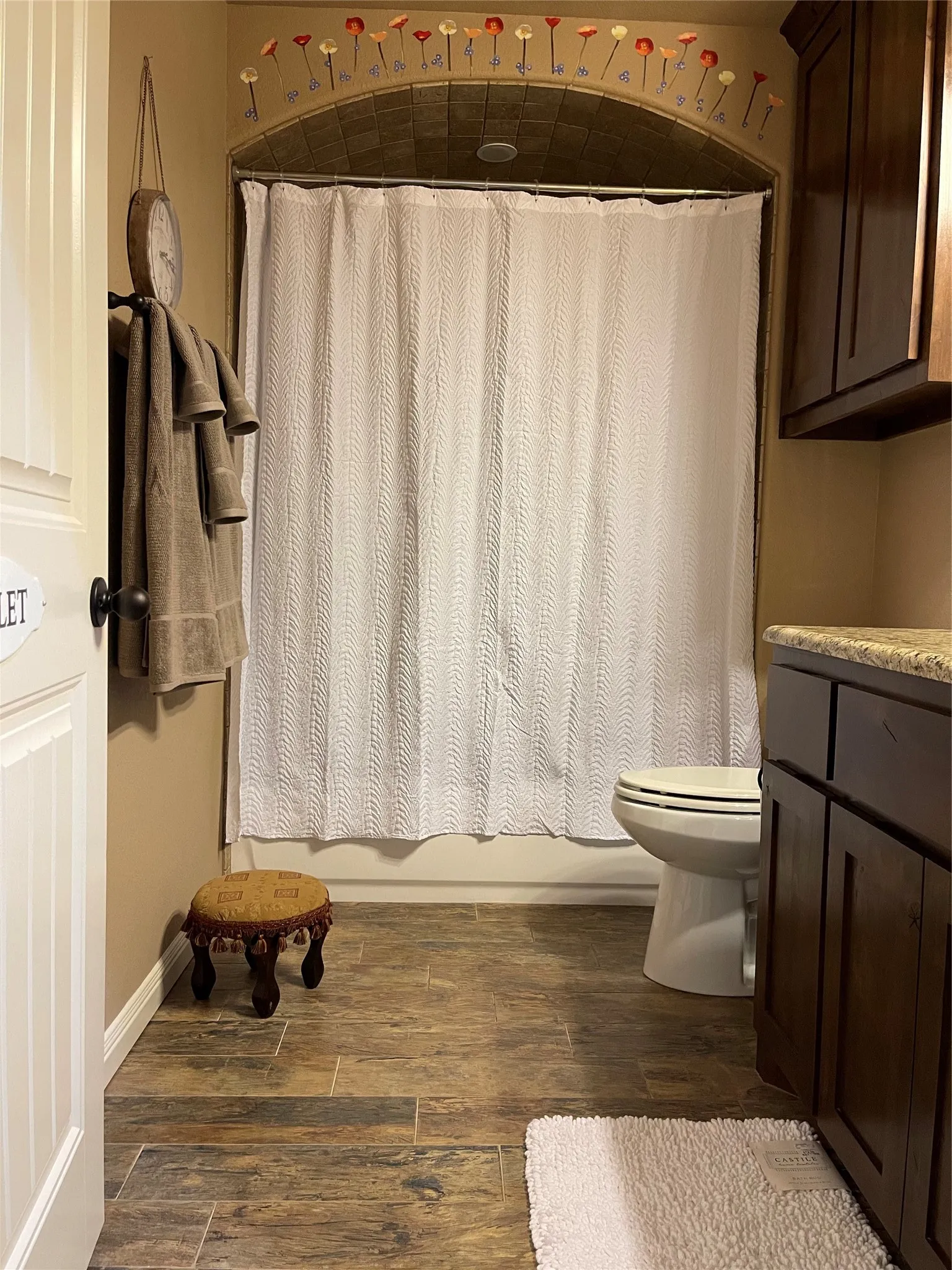 Arched shower alcove with tile surround, wood-finish flooring, dark wood cabinetry, and a stone-finish countertop