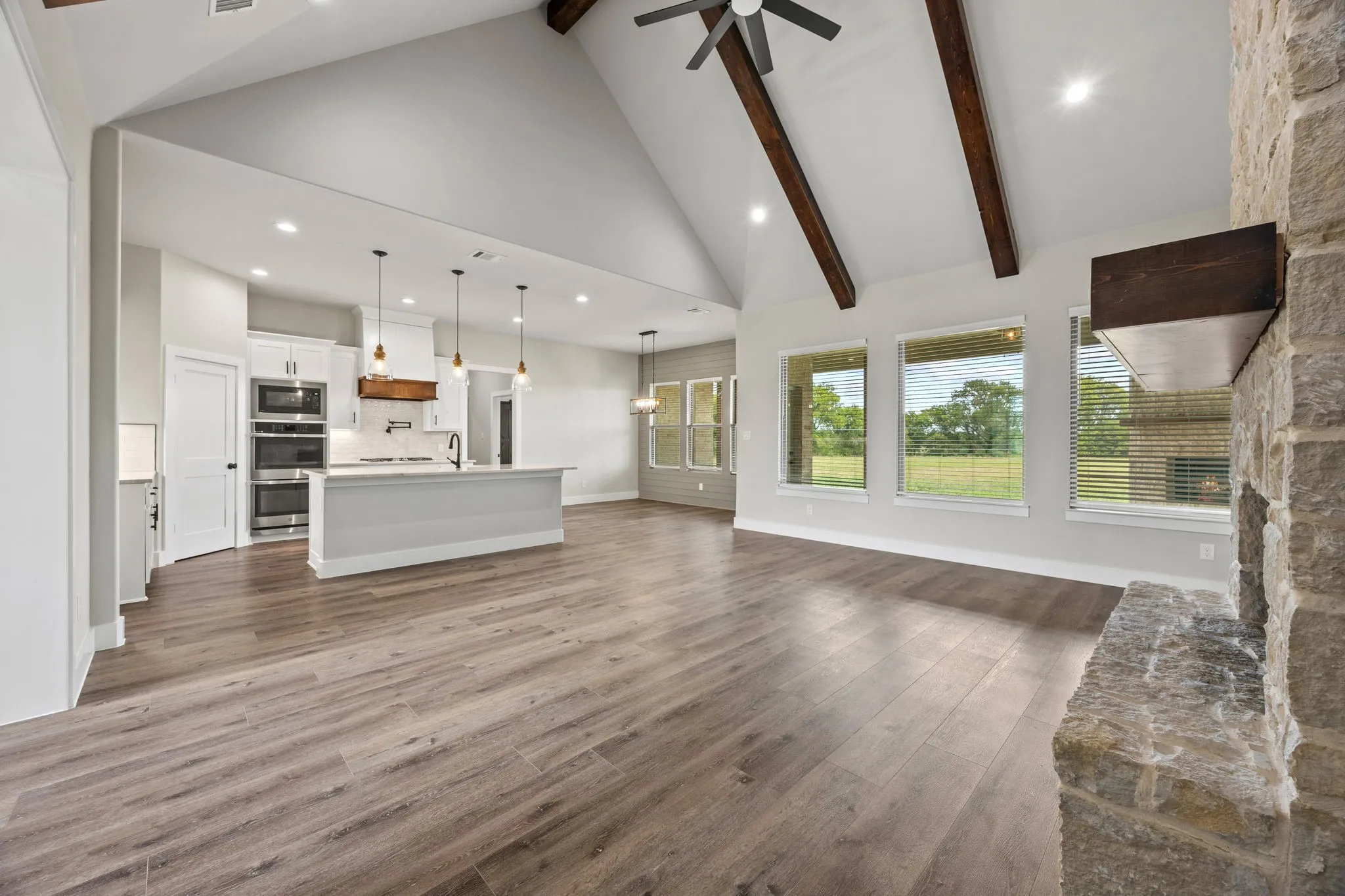 Vaulted ceiling with exposed wood beams, stone-faced fireplace with wood mantel, wood-finish flooring, white cabinetry with stainless steel appliances, and a kitchen island with pendant lighting