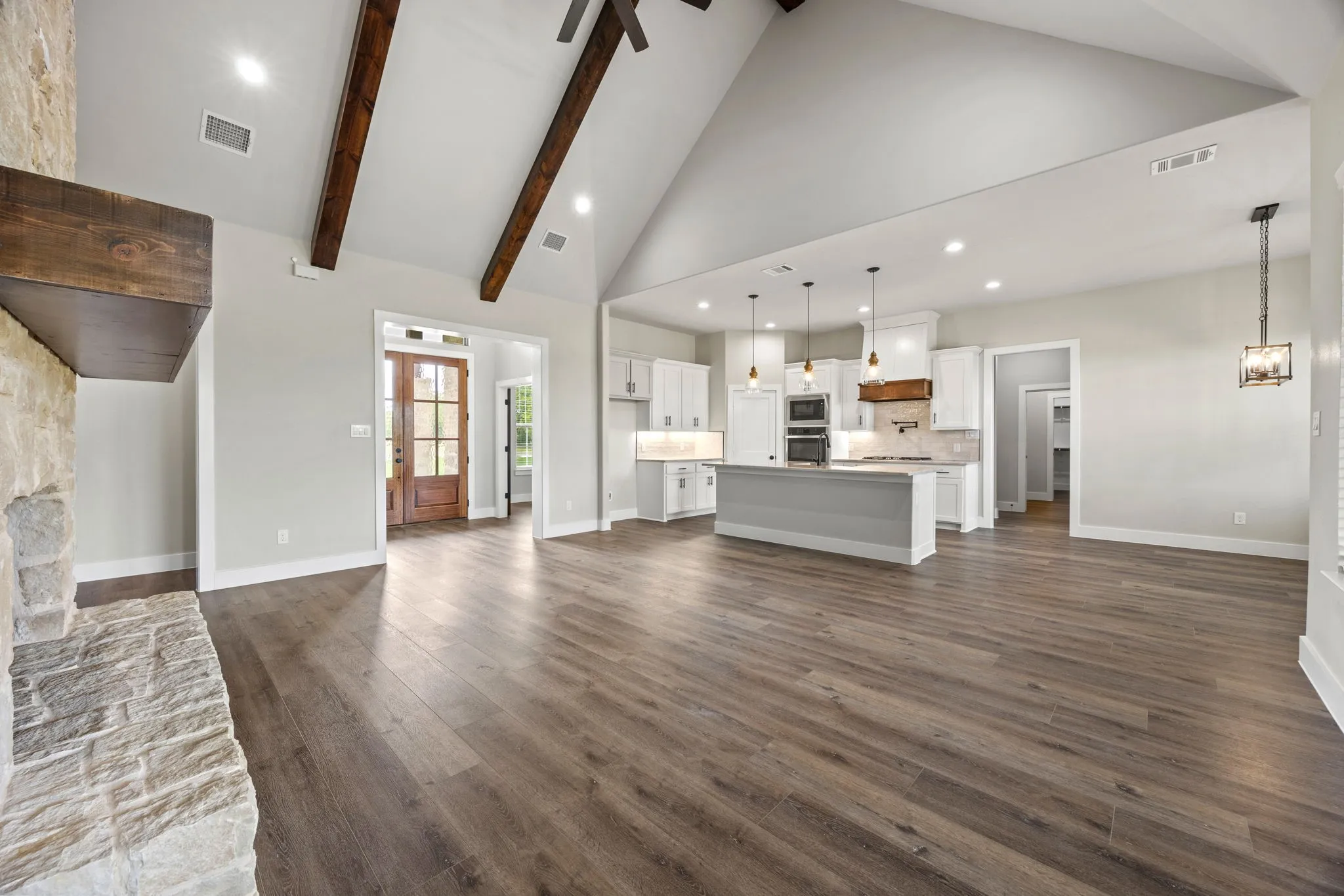 Vaulted ceiling with exposed wood beams, wood-finish flooring, stone fireplace with a wood mantle, kitchen island with pendant lighting, and white cabinetry