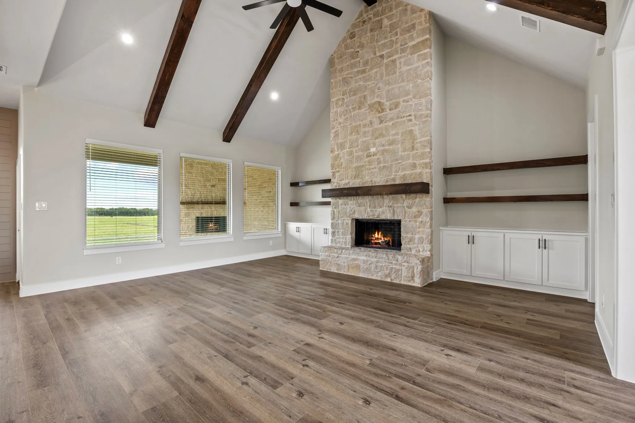 Vaulted ceiling with exposed wood beams, stone fireplace with a wood mantel, wood-finish flooring, and built-in cabinetry with open shelving