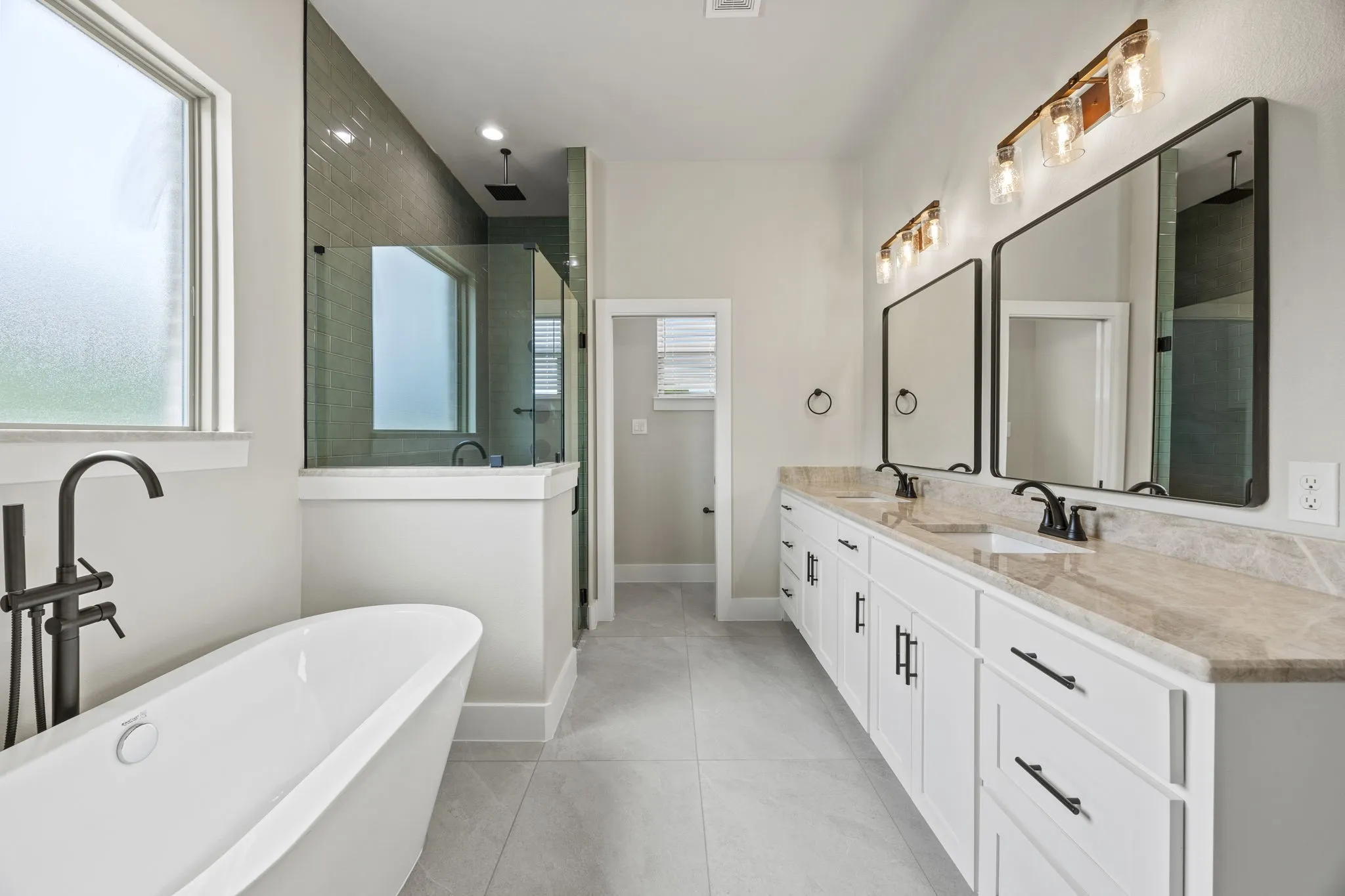Freestanding soaking tub with matte black fixtures, dual vanity with stone countertops and undermount sinks, separate glass-enclosed shower, and light gray tile flooring