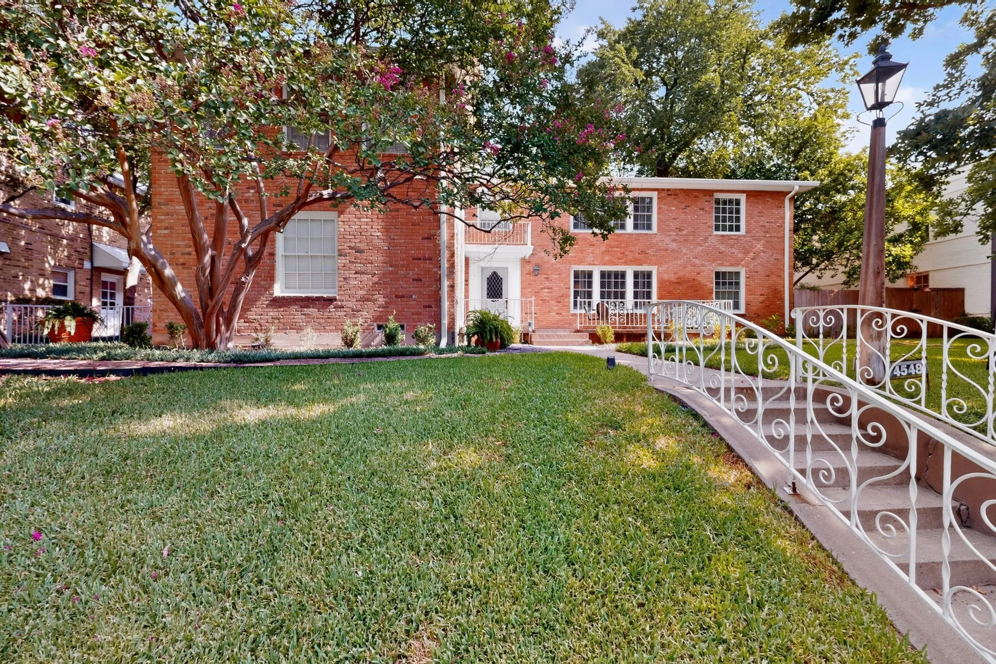 Brick exterior residence featuring multiple windows, a decorative metal railing, a prominent front entrance, and mature landscaping