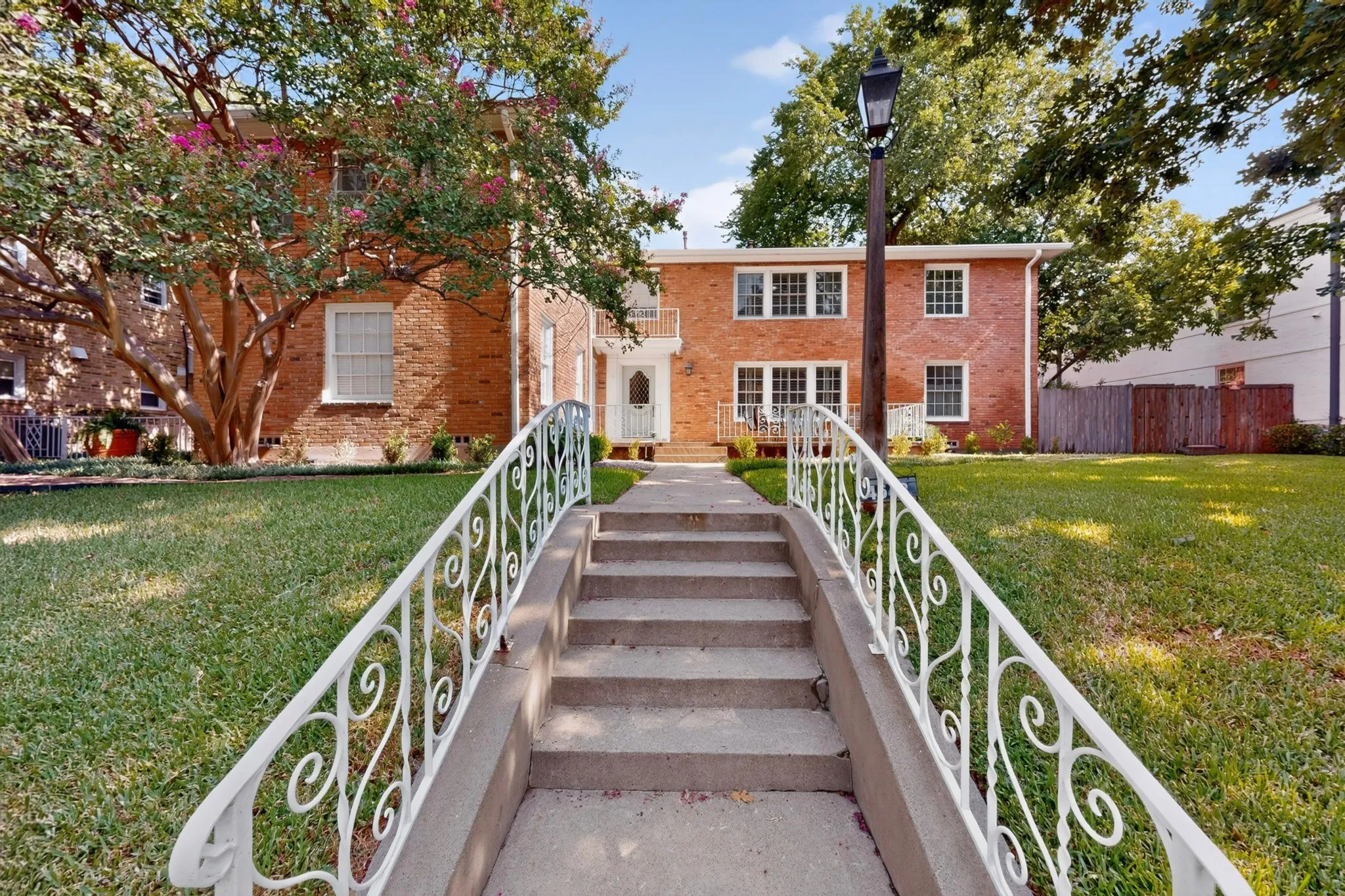 Brick exterior, concrete pathway with white decorative railings, mature trees, and a prominent street lamp