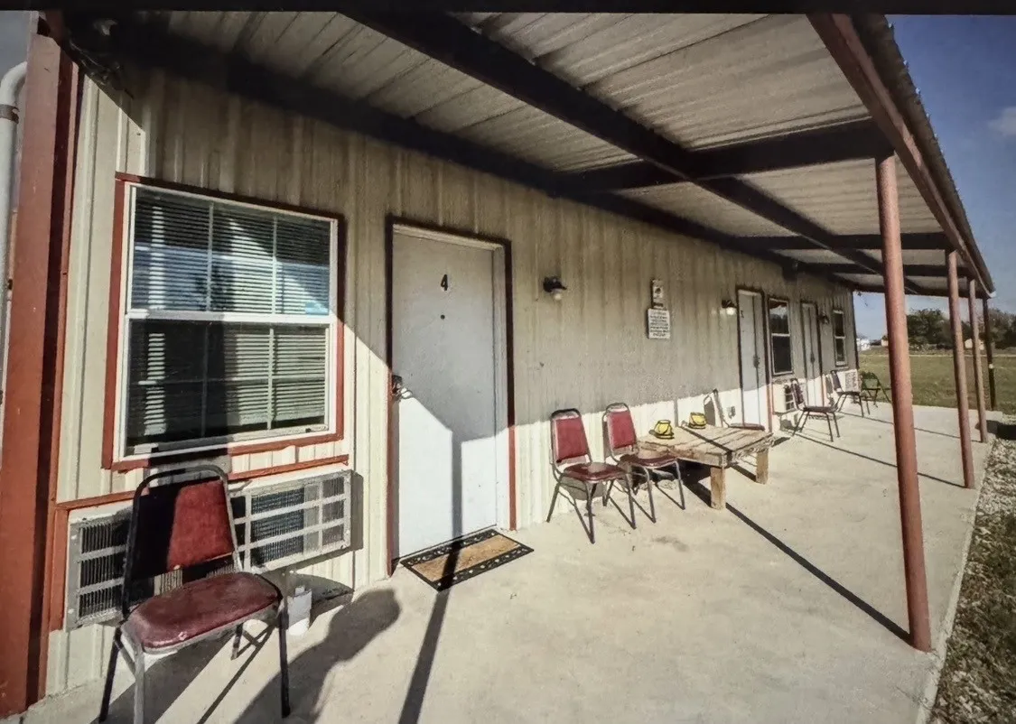 Metal-sided structure with multiple entry doors and windows, featuring a full-length covered porch supported by metal columns