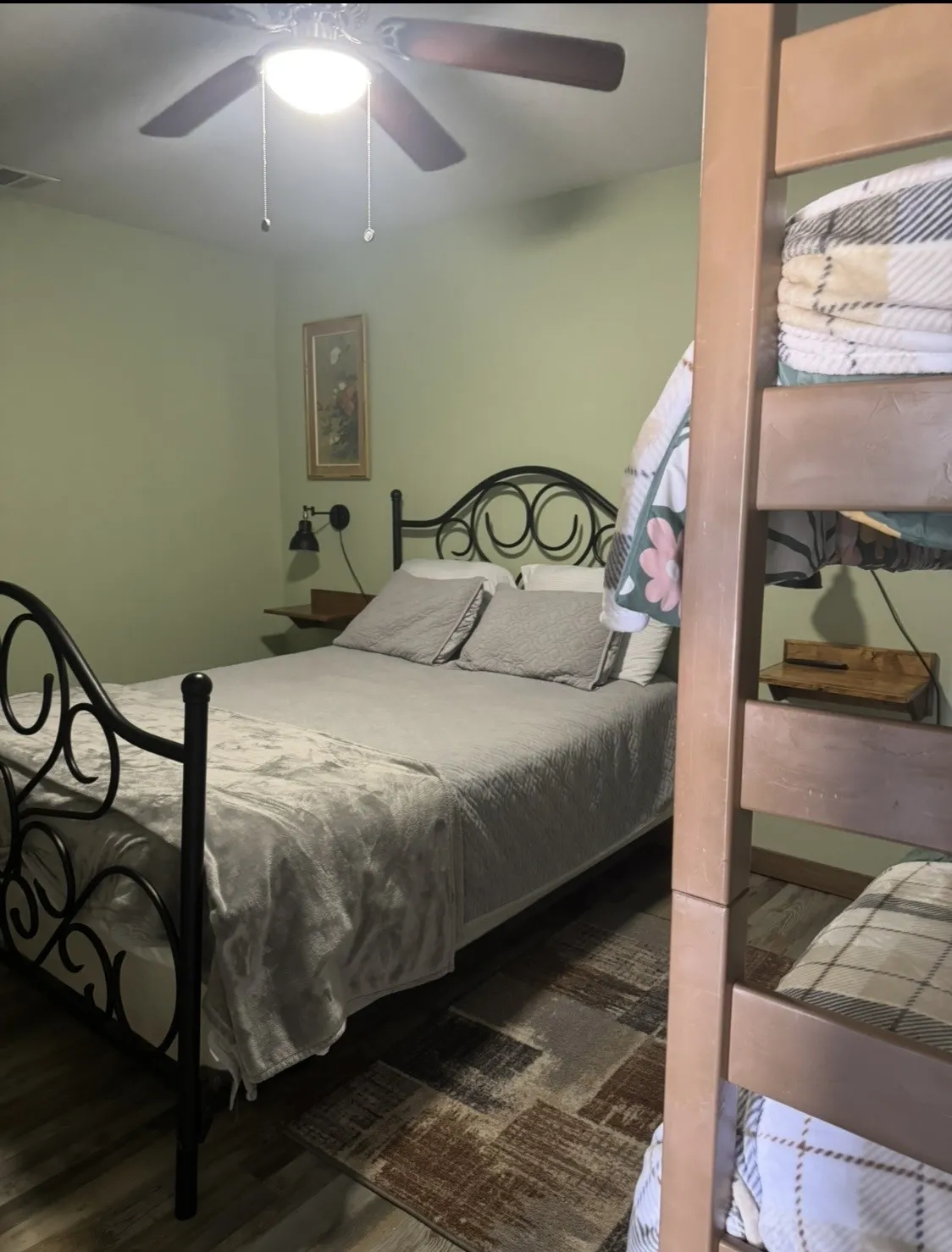 Wood-finish flooring with patterned rug, black metal bed frame, wall-mounted light fixture, and ceiling fan with integrated lighting