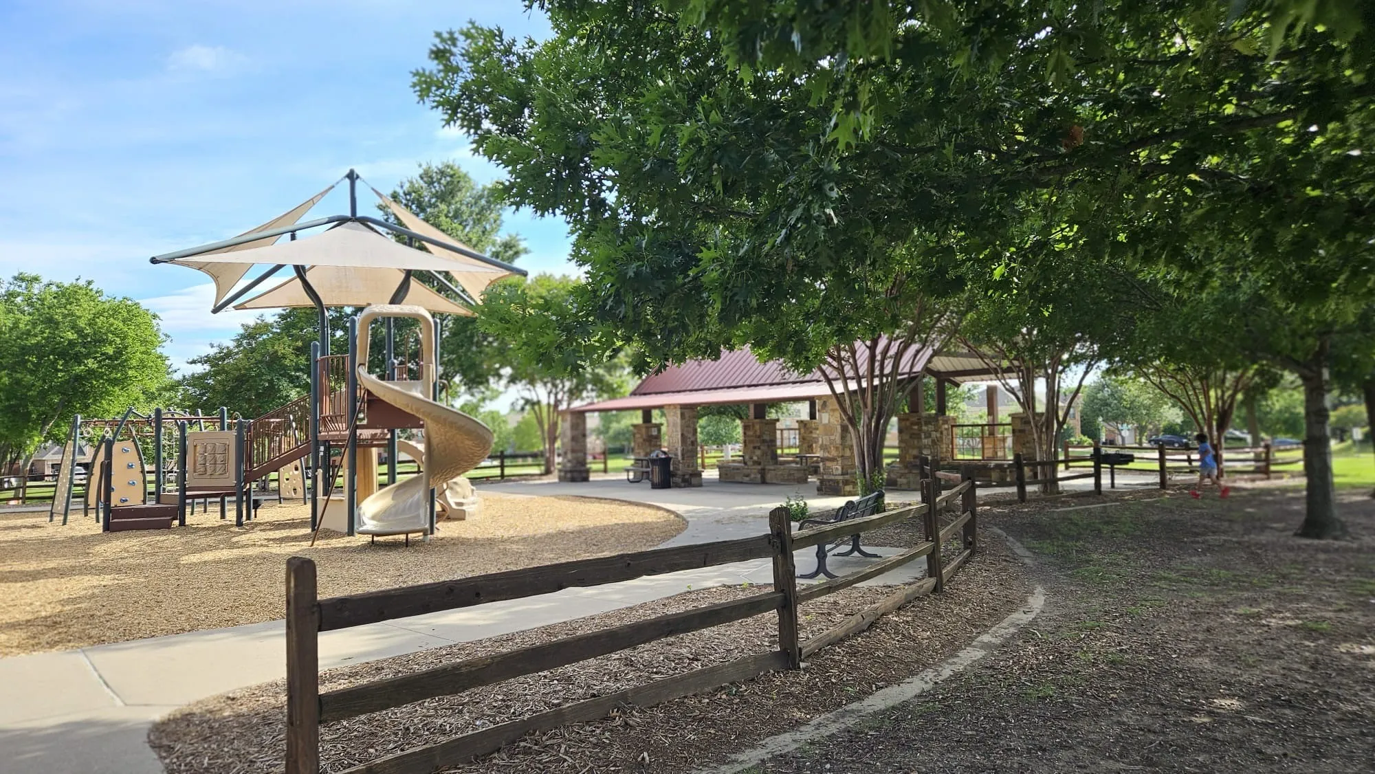 Park playground with shade structure, covered picnic area, paved pathways, and wood split rail fencing