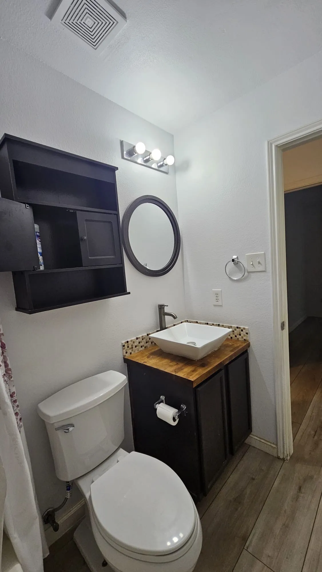 White vessel sink, wood-finish countertop, dark wood-finish vanity, wood-finish flooring, and round mirror