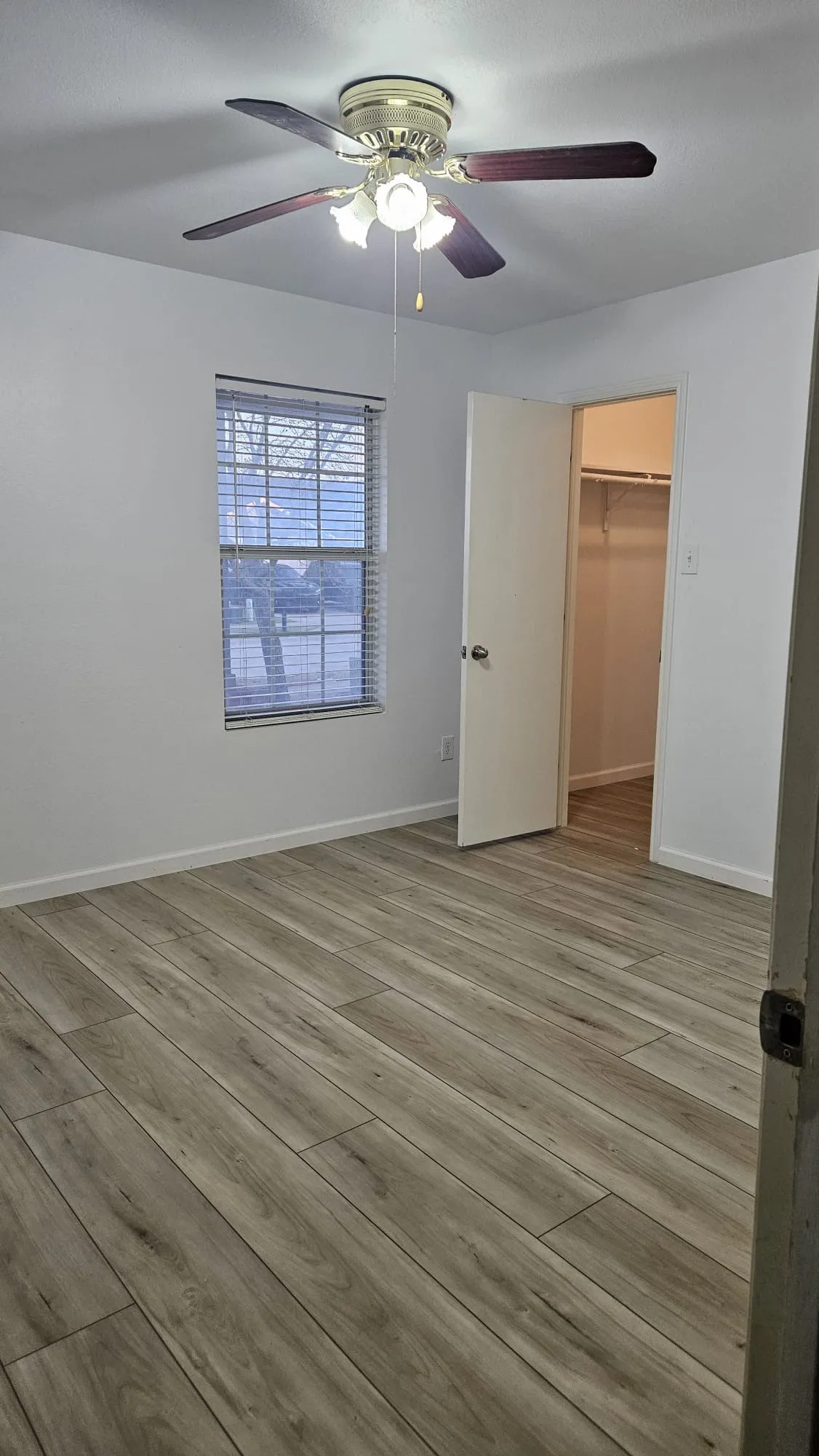 Wood-finish flooring throughout, bright white walls, window with horizontal blinds, and a ceiling fan with integrated lighting