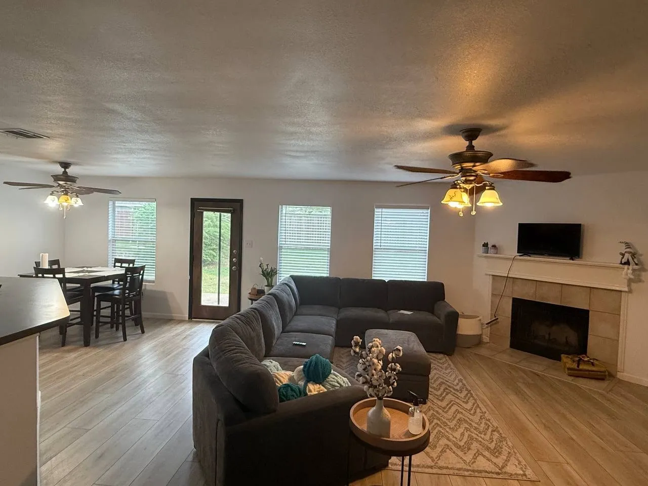 Open-concept living area featuring wood-finish flooring, a tiled fireplace with mantel, and two ceiling fans with integrated lighting