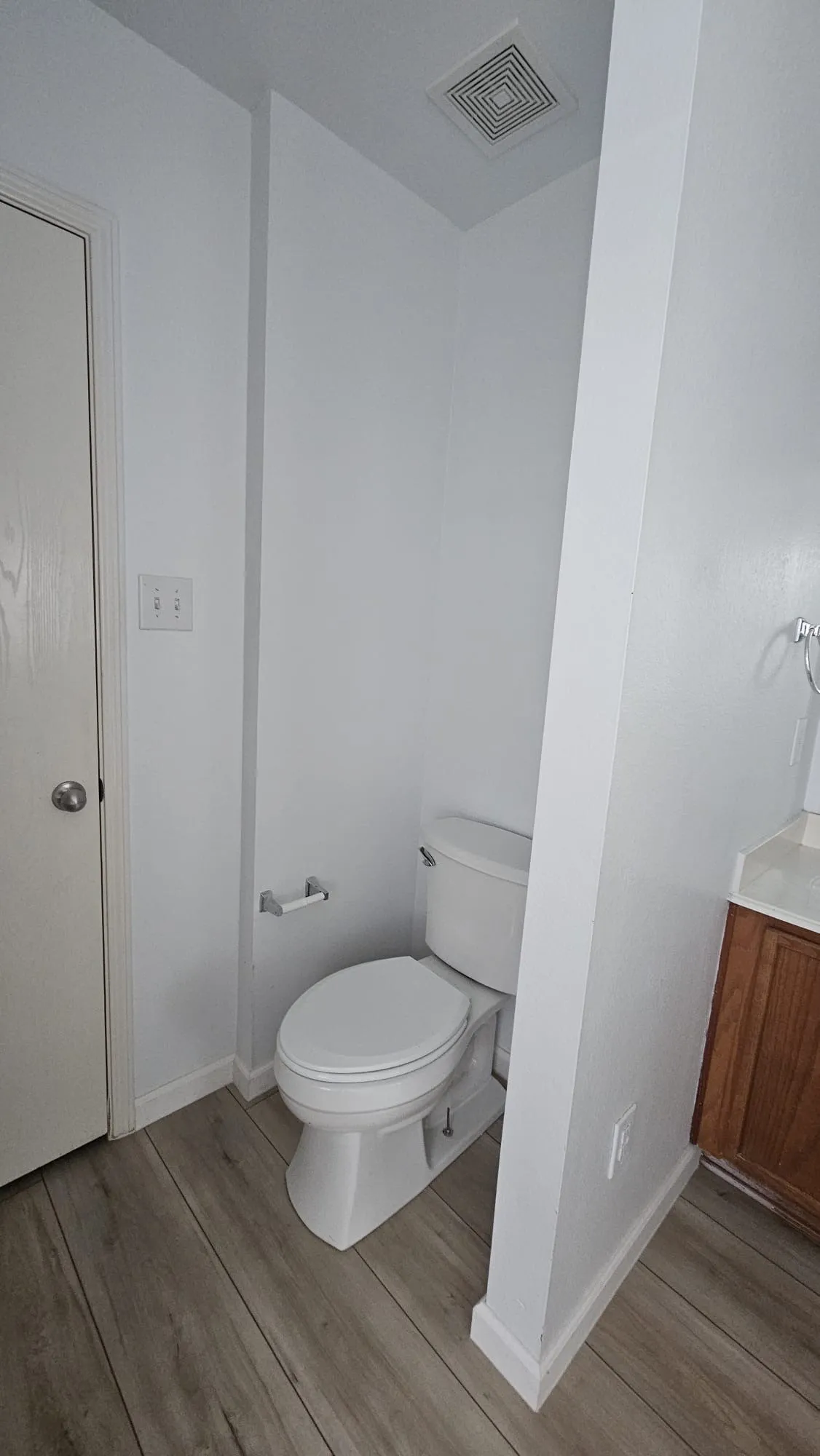 White toilet with wood-finish flooring, white walls, and a recessed ceiling vent