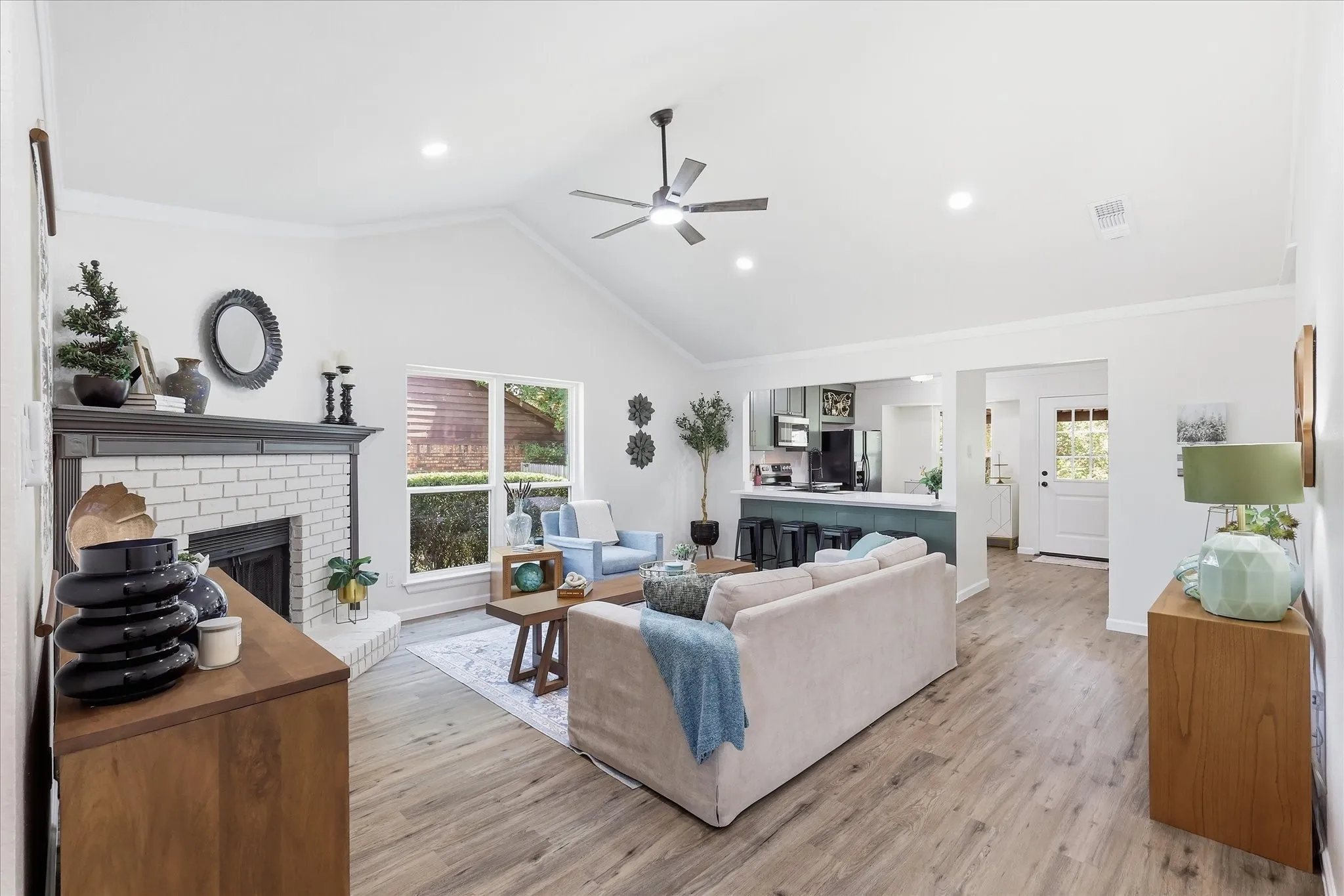Vaulted ceiling with recessed lighting, wood-finish flooring, white brick fireplace with dark mantel, and a kitchen pass-through