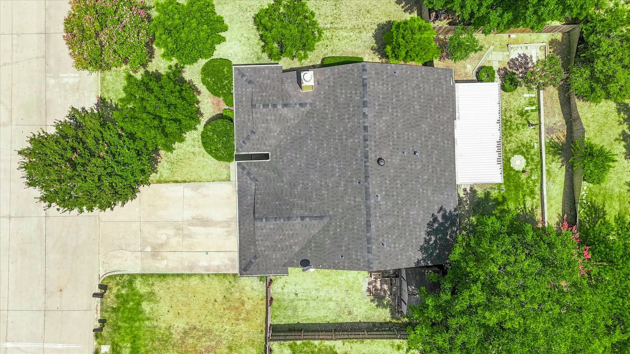 Asphalt shingle roof, concrete driveway, concrete patio, perimeter fencing, and mature landscaping