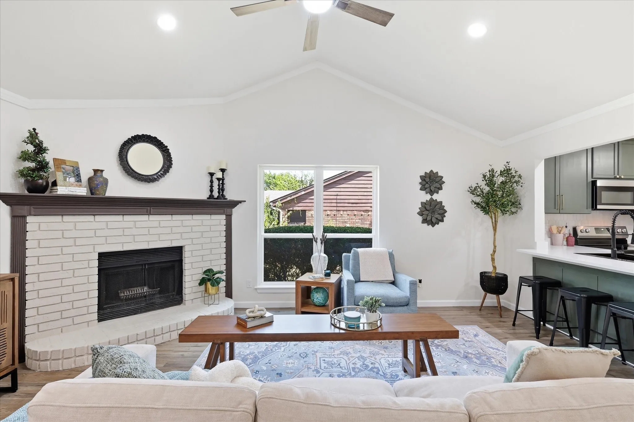 Vaulted ceilings, a white brick fireplace with a dark wood mantel, wood-finish flooring, and a ceiling fan with integrated lighting