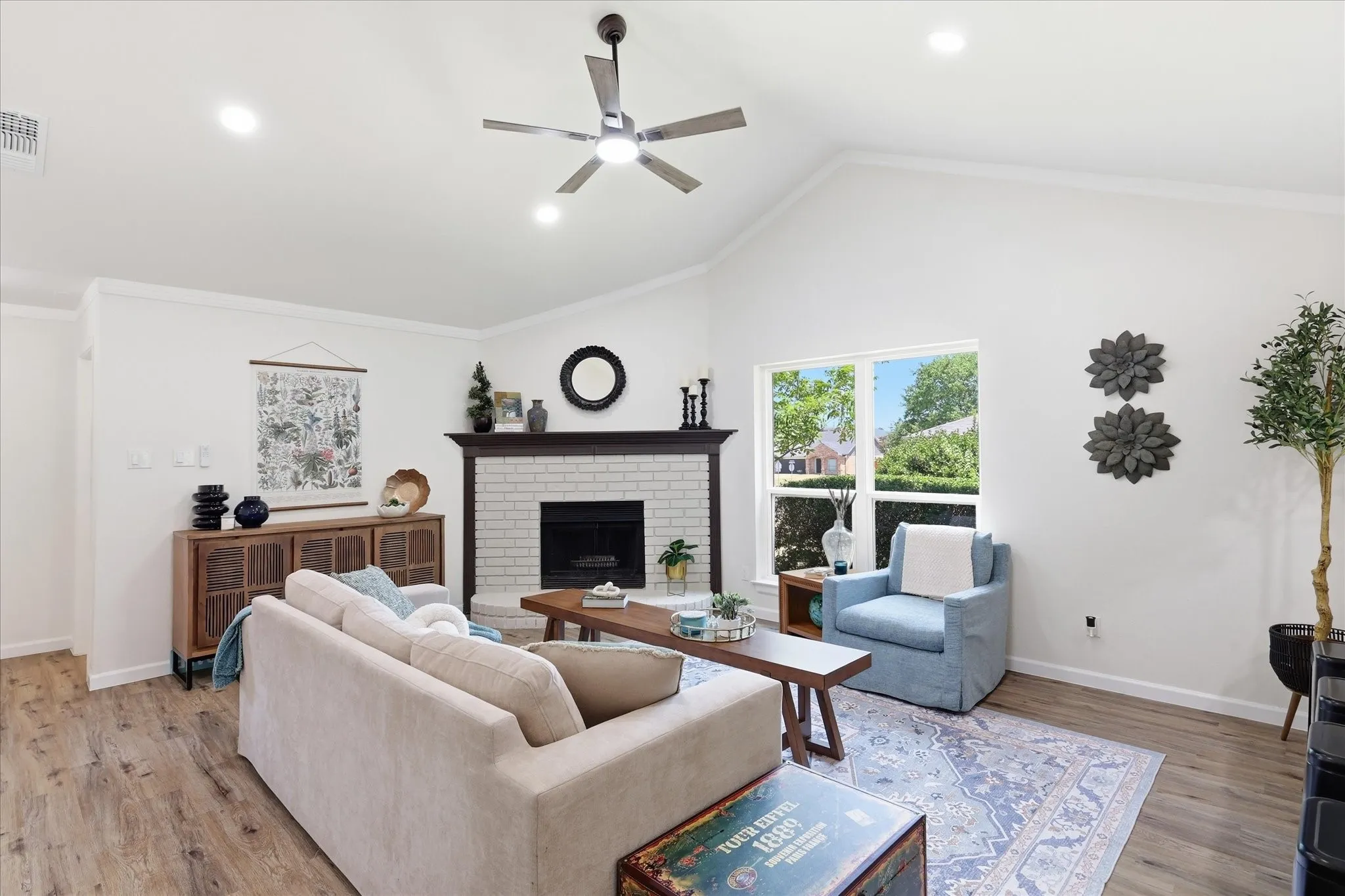 Vaulted ceiling, wood-finish flooring, white brick fireplace, window with exterior landscaping, and a ceiling fan with integrated lighting