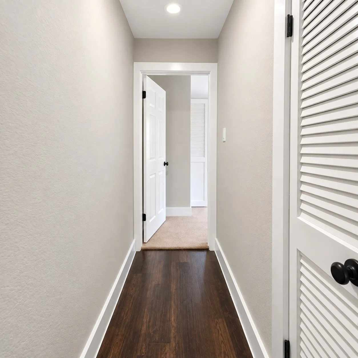 Hallway featuring wood-finish flooring, recessed lighting, white trim, a white paneled door with black hardware, and a white louvered door with black hardware