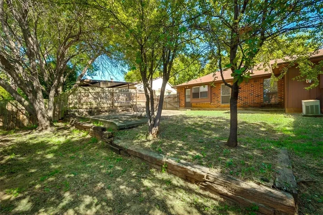Expansive backyard featuring a concrete patio, mature trees, wood-finish retaining walls, and a wood fence