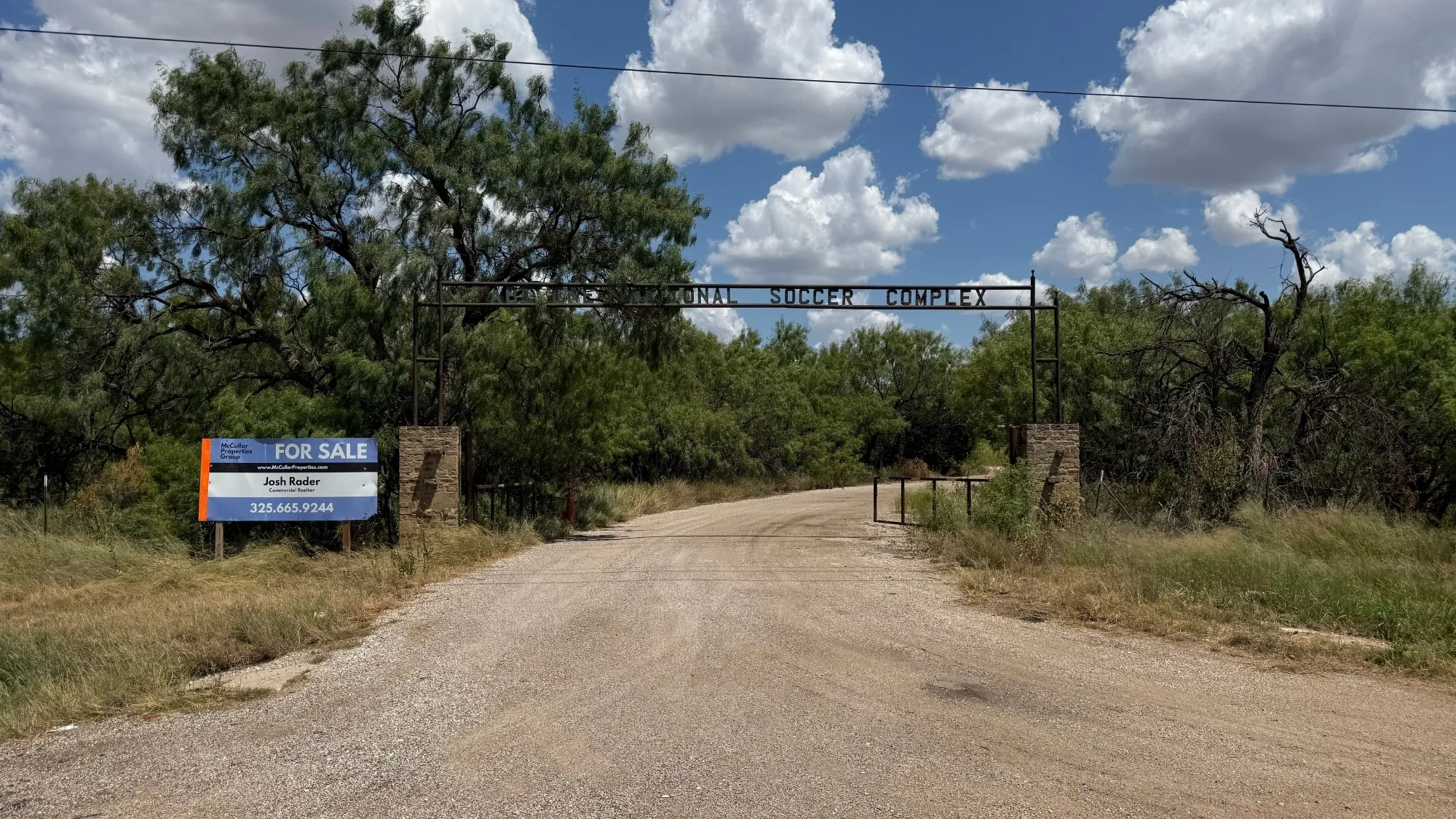 Gravel road entrance with a metal archway and stone pillars, surrounded by mature trees and natural landscaping