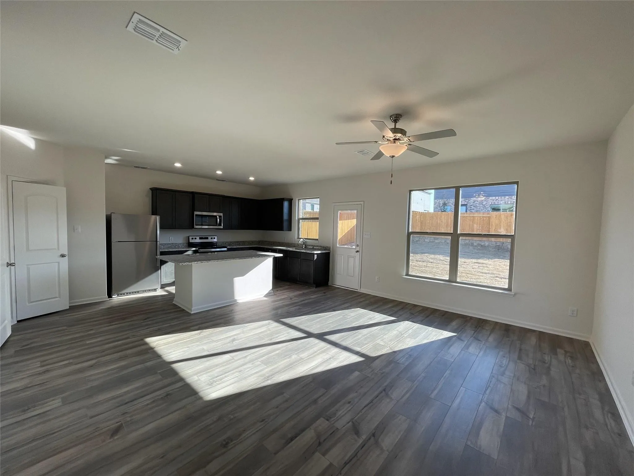 Open-concept living area featuring wood-finish flooring, a kitchen island with a countertop, stainless steel appliances, dark cabinetry, and a ceiling fan with integrated lighting