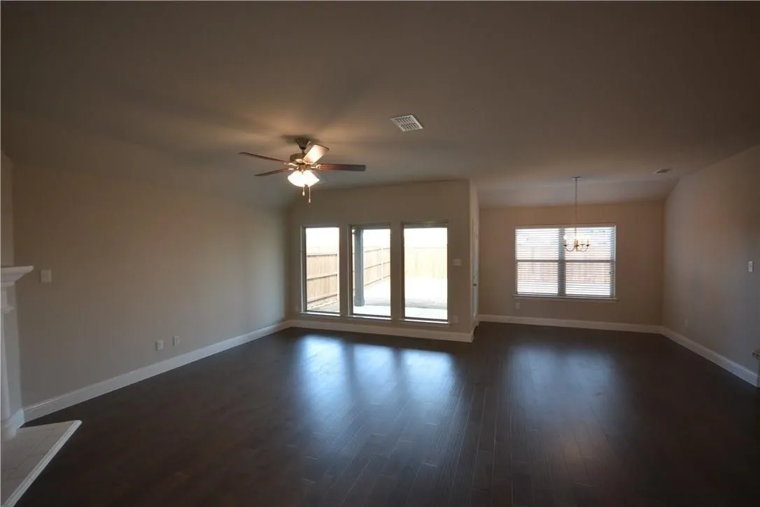 Spacious open-concept interior featuring dark wood-finish flooring, multiple windows, a ceiling fan with integrated lighting, and a decorative chandelier