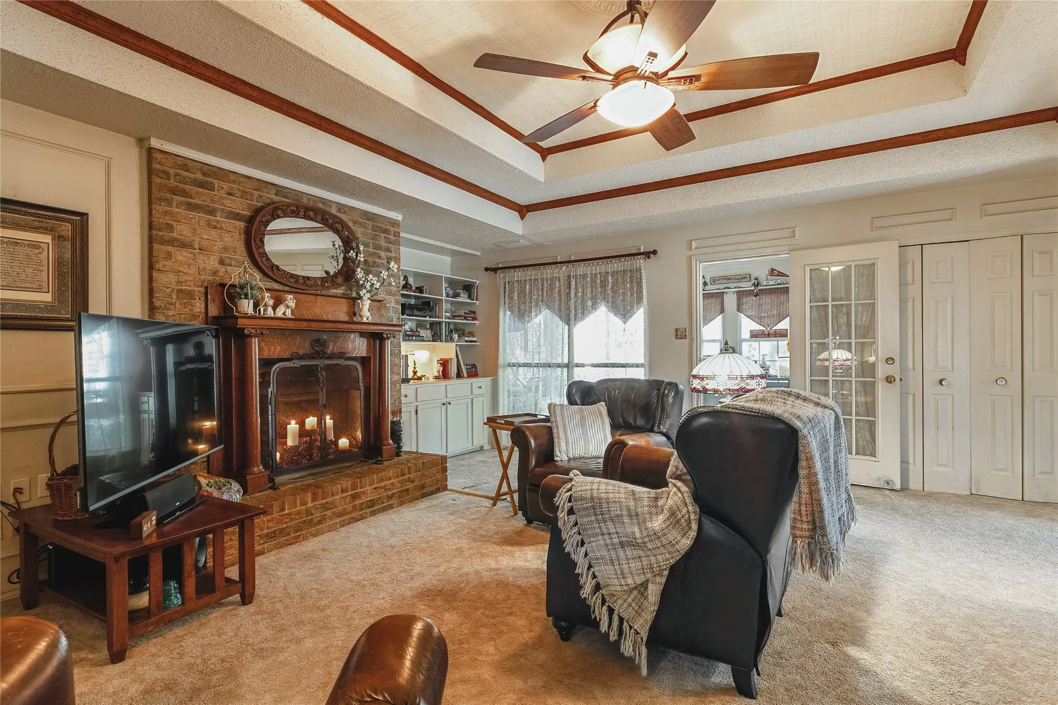 Tray ceiling with wood-finish trim, brick fireplace with wood mantel, built-in shelving, glass-paned doors, and carpet flooring